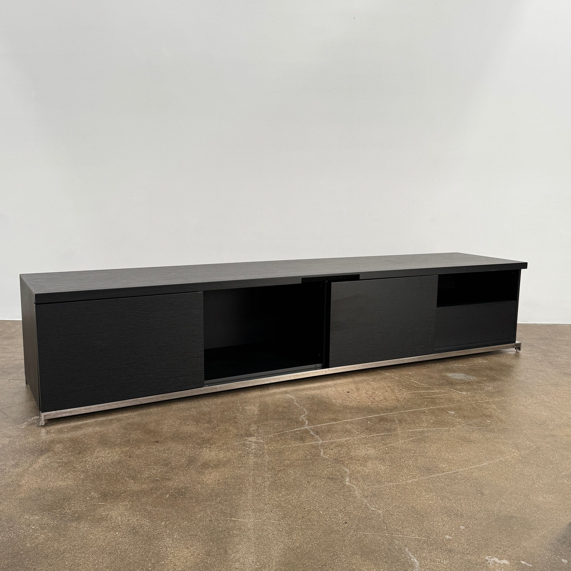 The Maxalto Mida Console by Maxalto is a modern black sideboard TV stand with three closed cabinets and two open shelves, shown on a concrete floor against a plain white background.