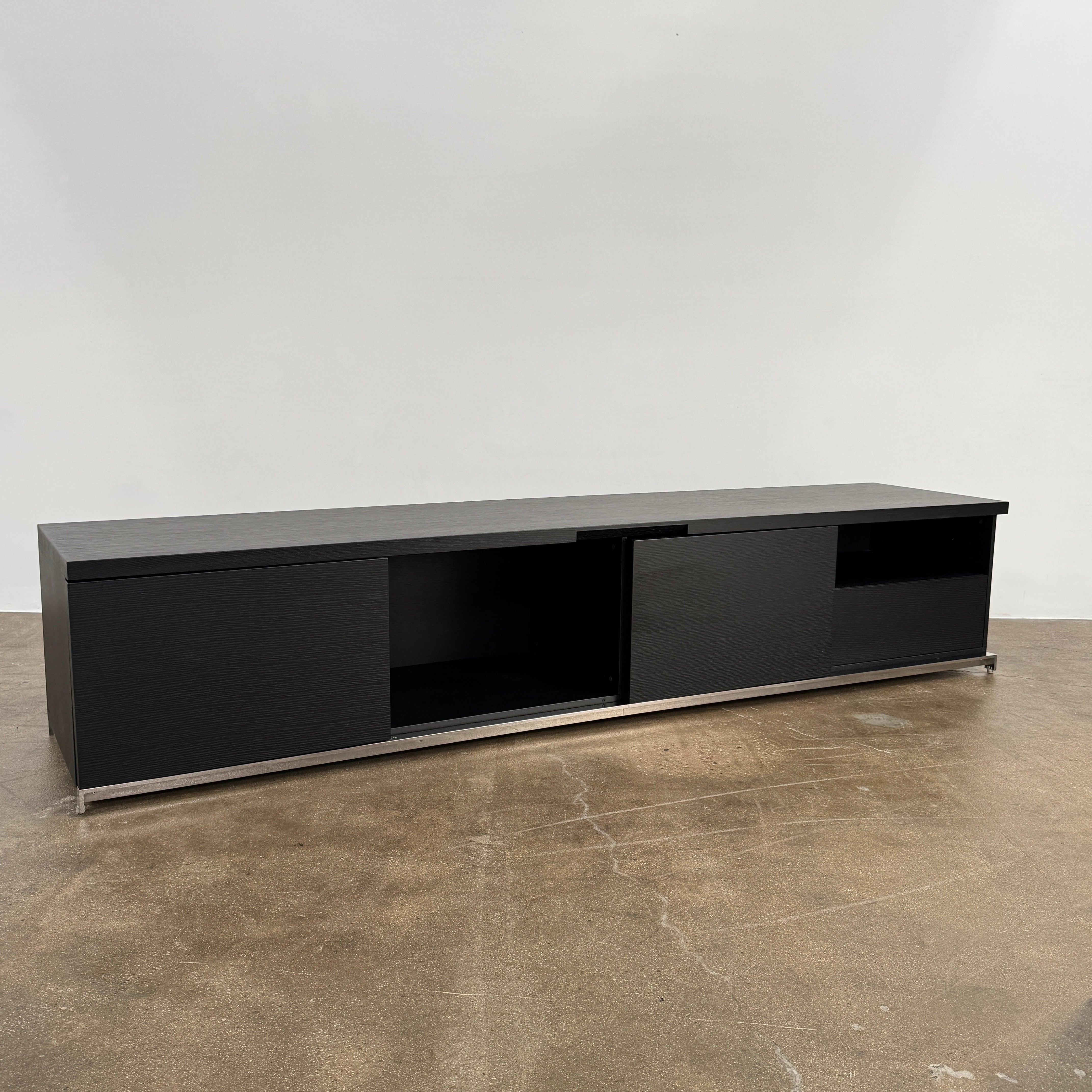 The Maxalto Mida Console by Maxalto is a modern black sideboard TV stand with three closed cabinets and two open shelves, shown on a concrete floor against a plain white background.