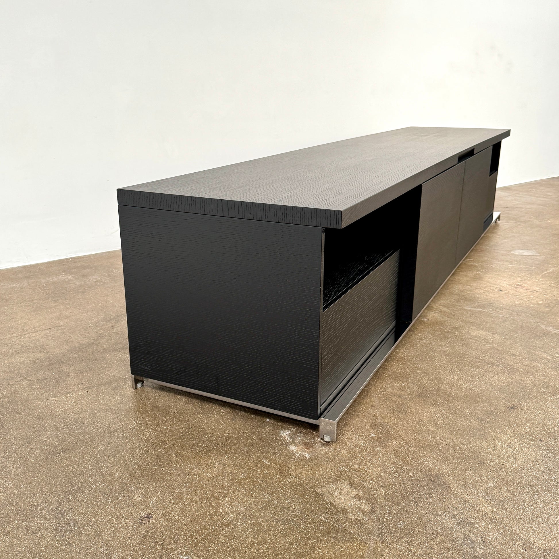 The Maxalto Mida Console by Maxalto is a modern black sideboard TV stand with three closed cabinets and two open shelves, shown on a concrete floor against a plain white background.
