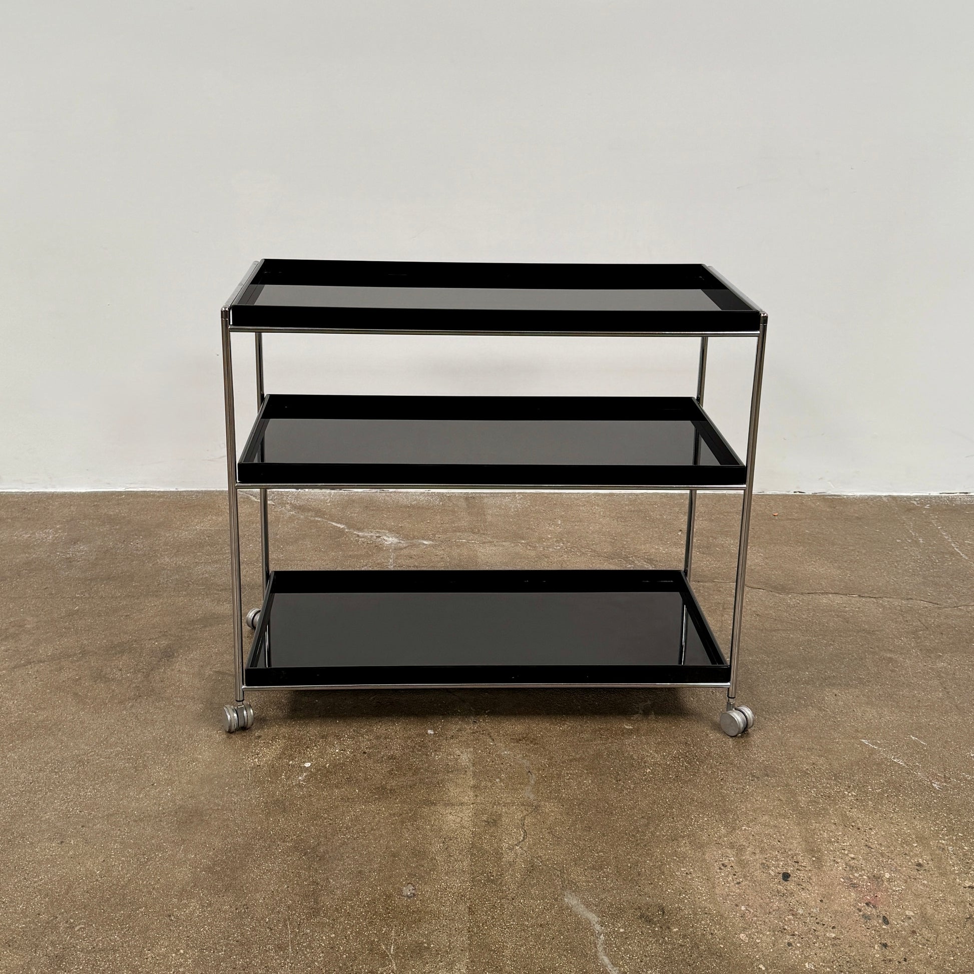 The Kartell Trays Trolly by Kartell features three black metal tiers, glass shelves, and four wheels, displayed on a plain white background.