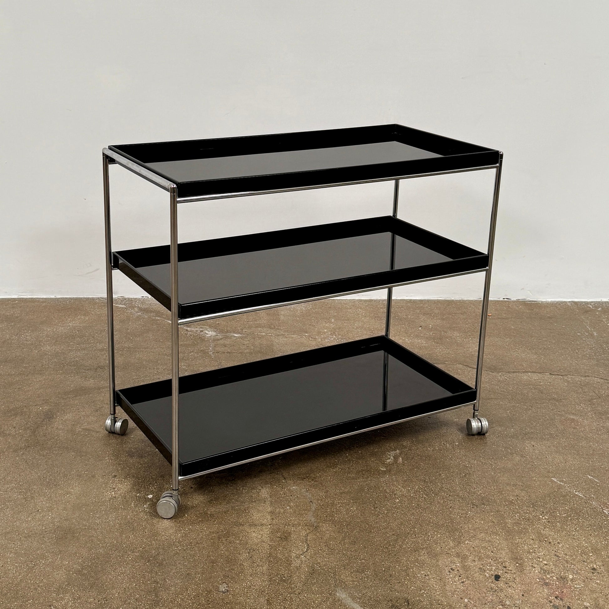 The Kartell Trays Trolly by Kartell features three black metal tiers, glass shelves, and four wheels, displayed on a plain white background.