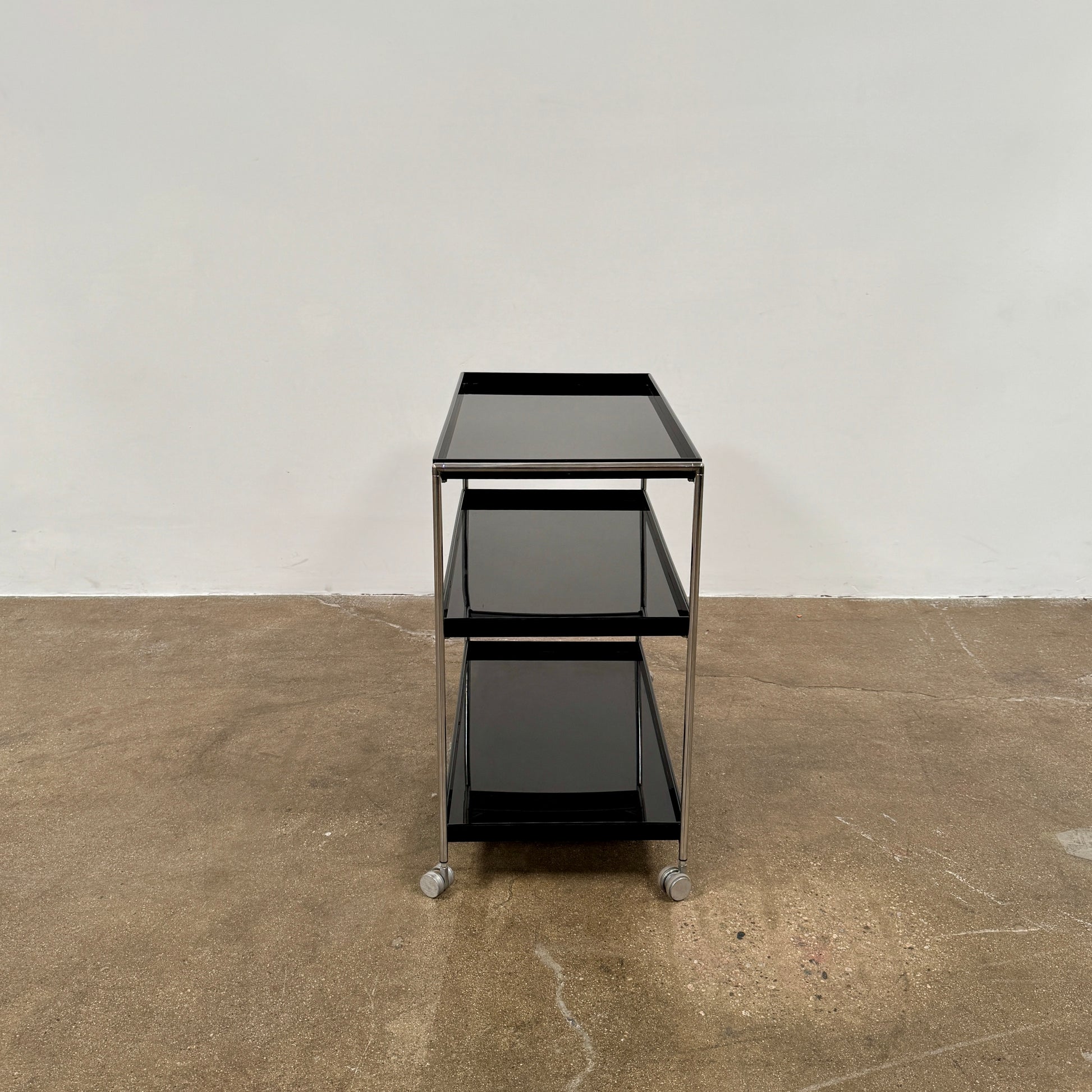 The Kartell Trays Trolly by Kartell features three black metal tiers, glass shelves, and four wheels, displayed on a plain white background.