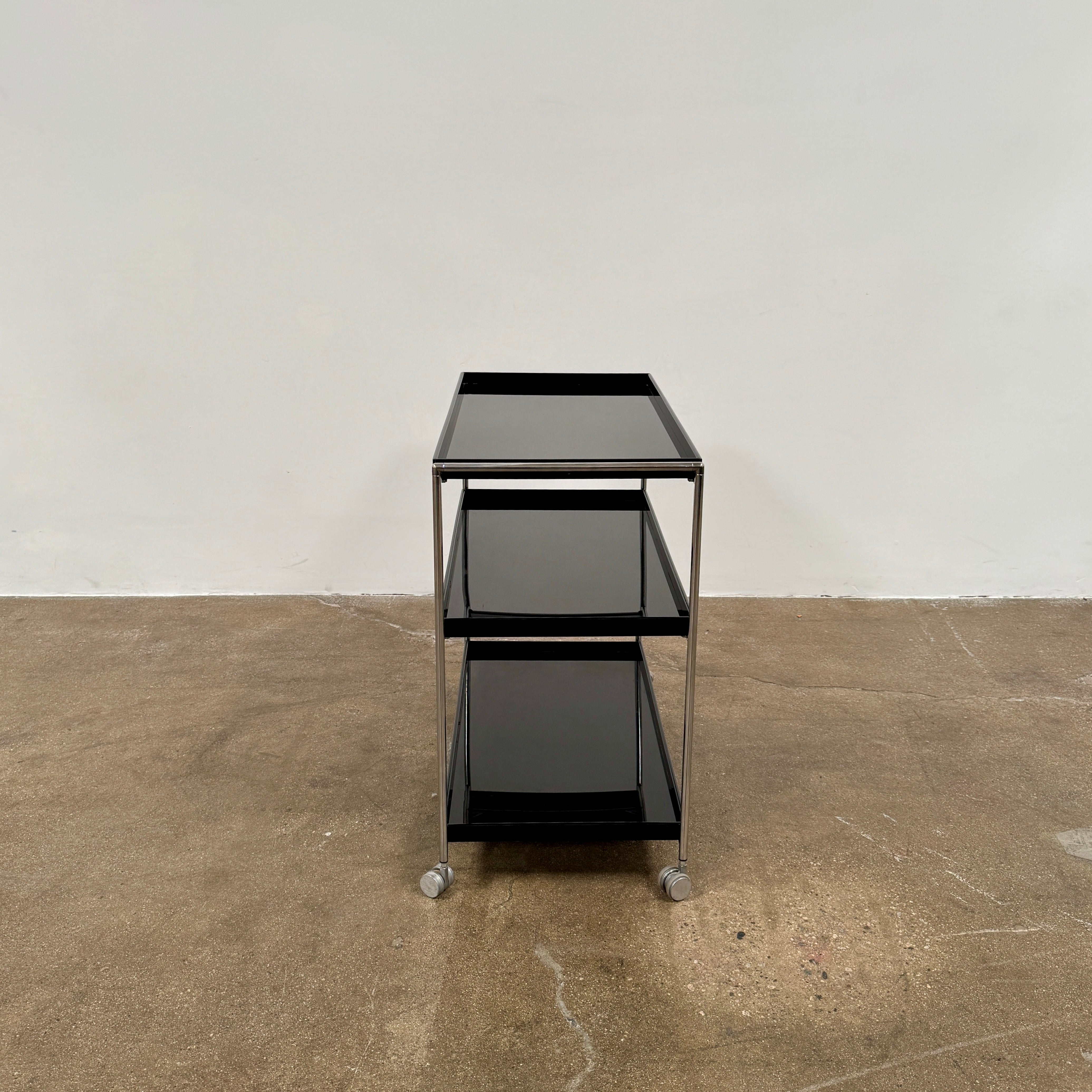 The Kartell Trays Trolly by Kartell features three black metal tiers, glass shelves, and four wheels, displayed on a plain white background.