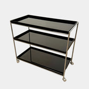 The Kartell Trays Trolly by Kartell features three black metal tiers, glass shelves, and four wheels, displayed on a plain white background.