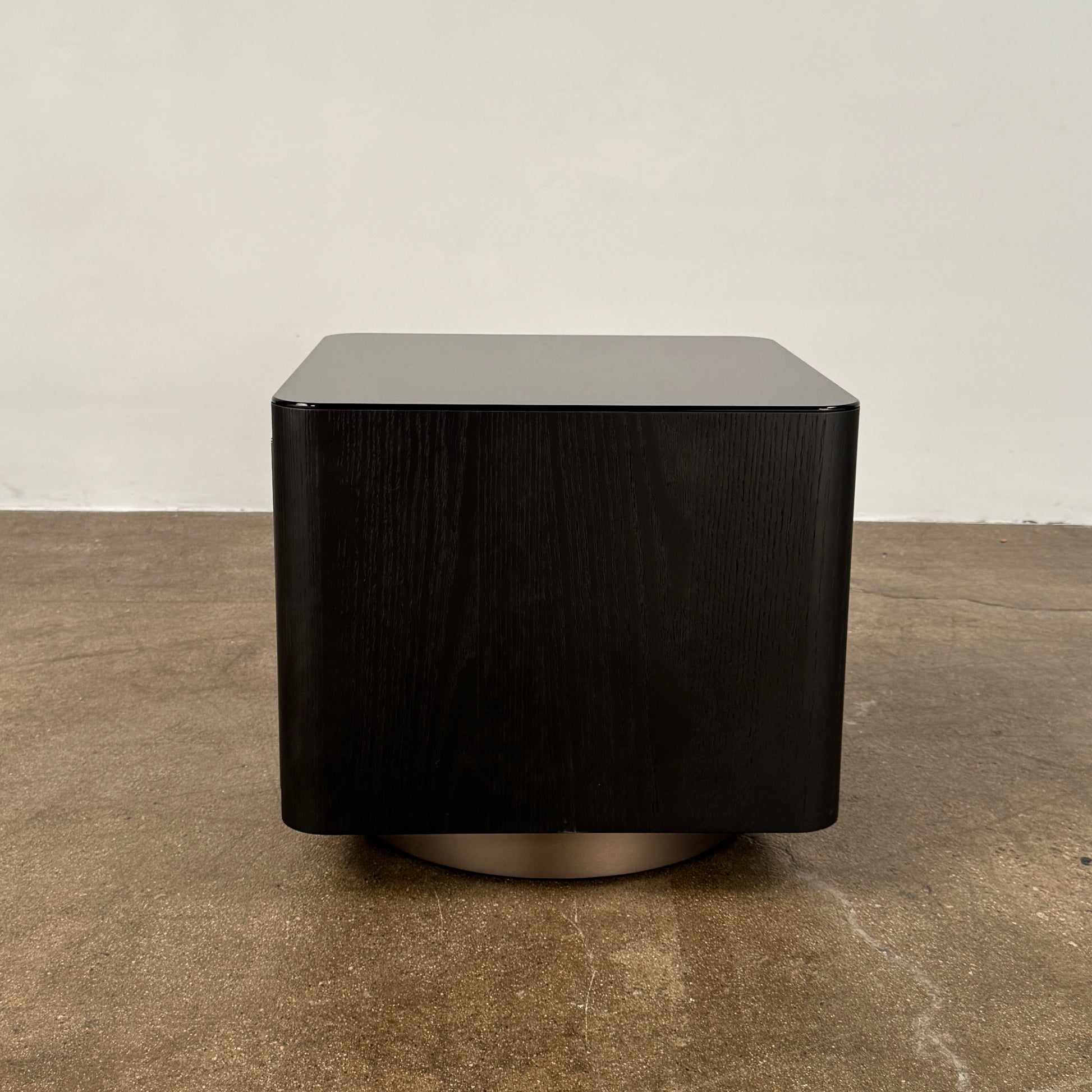 The Minotti Milton Side Table by Minotti features a modern, dark wooden cube design with an open center and a circular base, displayed on a polished concrete floor against a plain white wall.