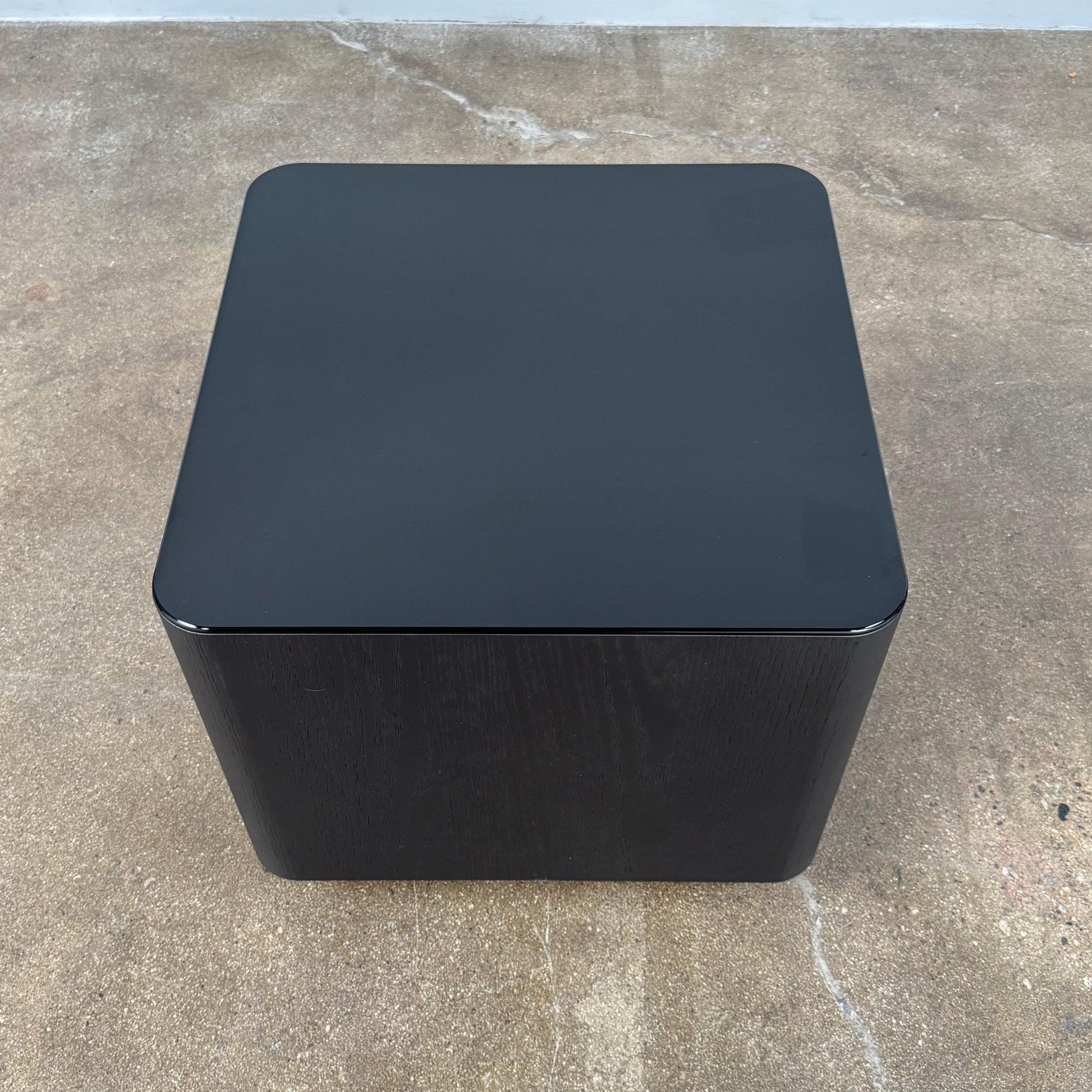 The Minotti Milton Side Table by Minotti features a modern, dark wooden cube design with an open center and a circular base, displayed on a polished concrete floor against a plain white wall.