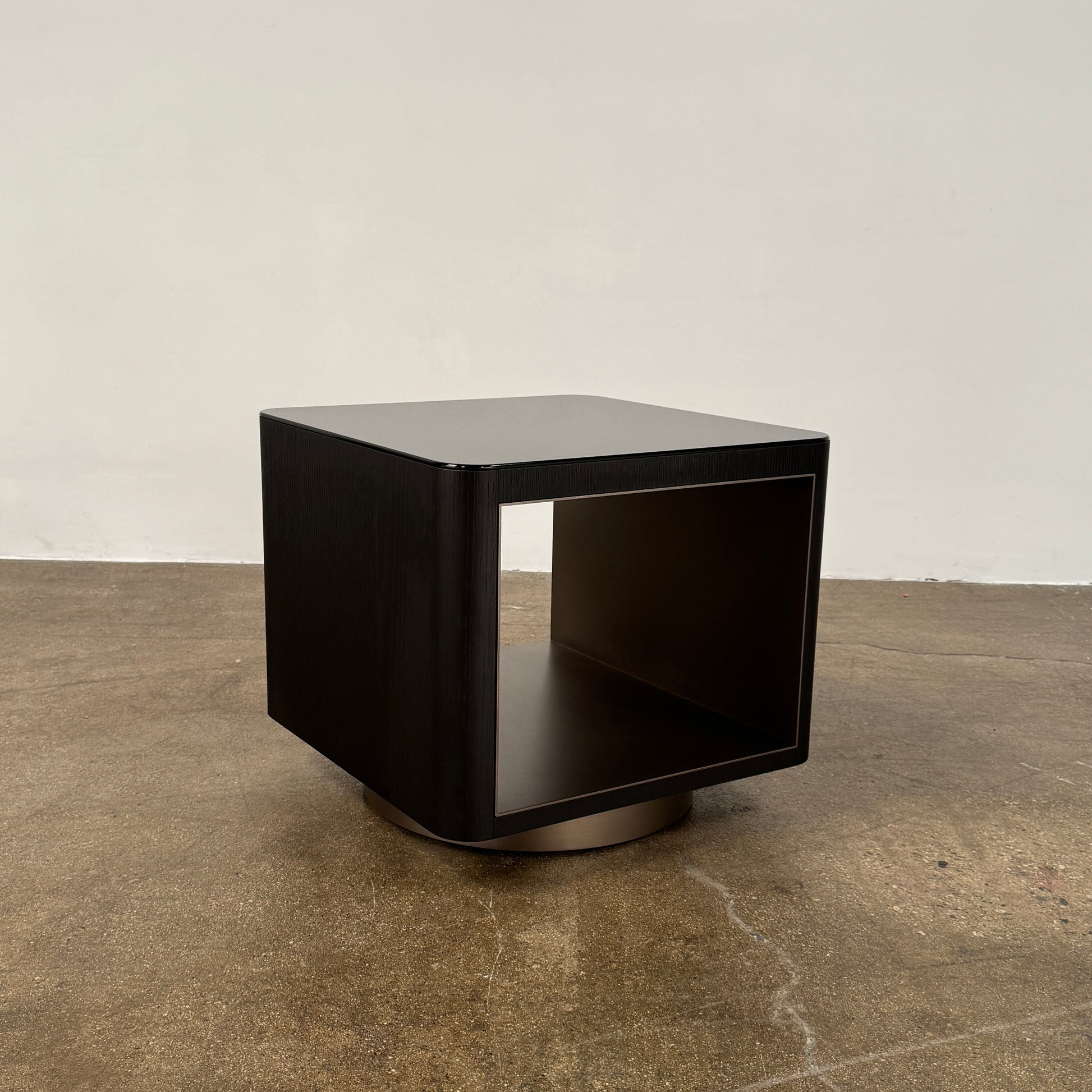 The Minotti Milton Side Table by Minotti features a modern, dark wooden cube design with an open center and a circular base, displayed on a polished concrete floor against a plain white wall.