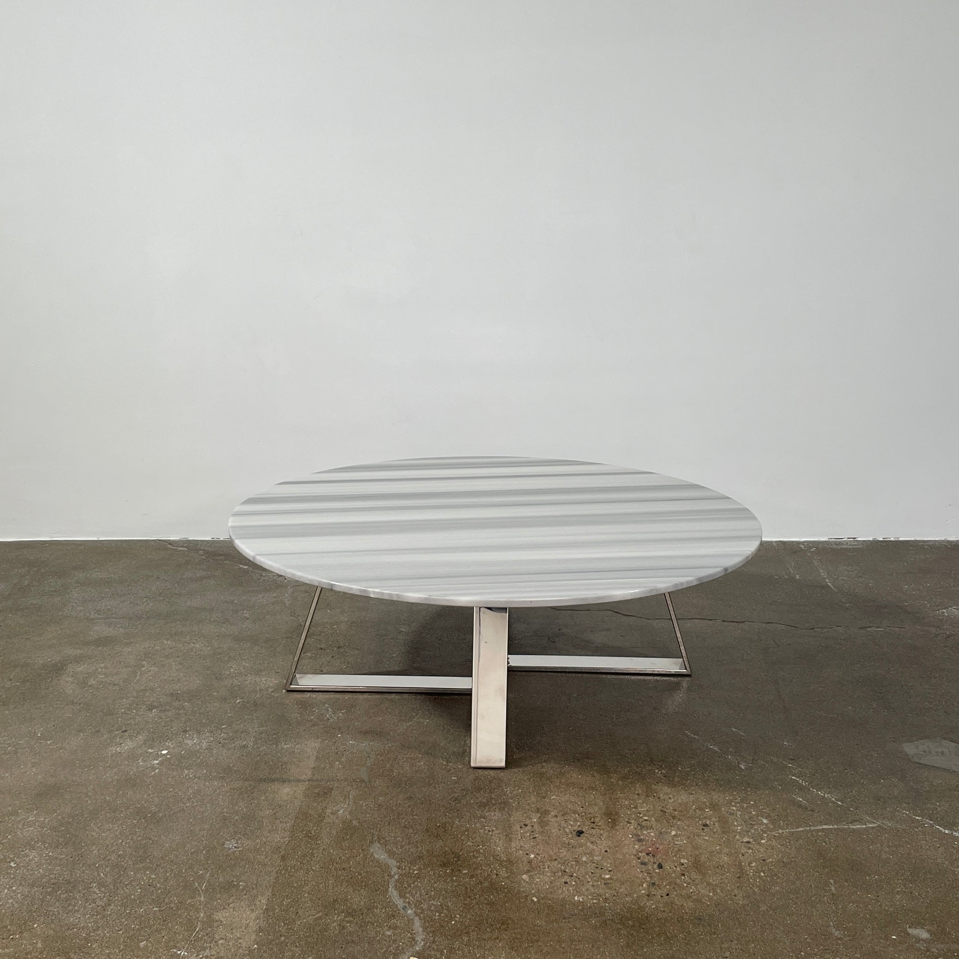 The Lievo Home Modern Studio Ingrid Coffee Table has a glossy marble top with white and gray stripes and a polished stainless steel base, shown on a bare concrete floor against a plain white wall.