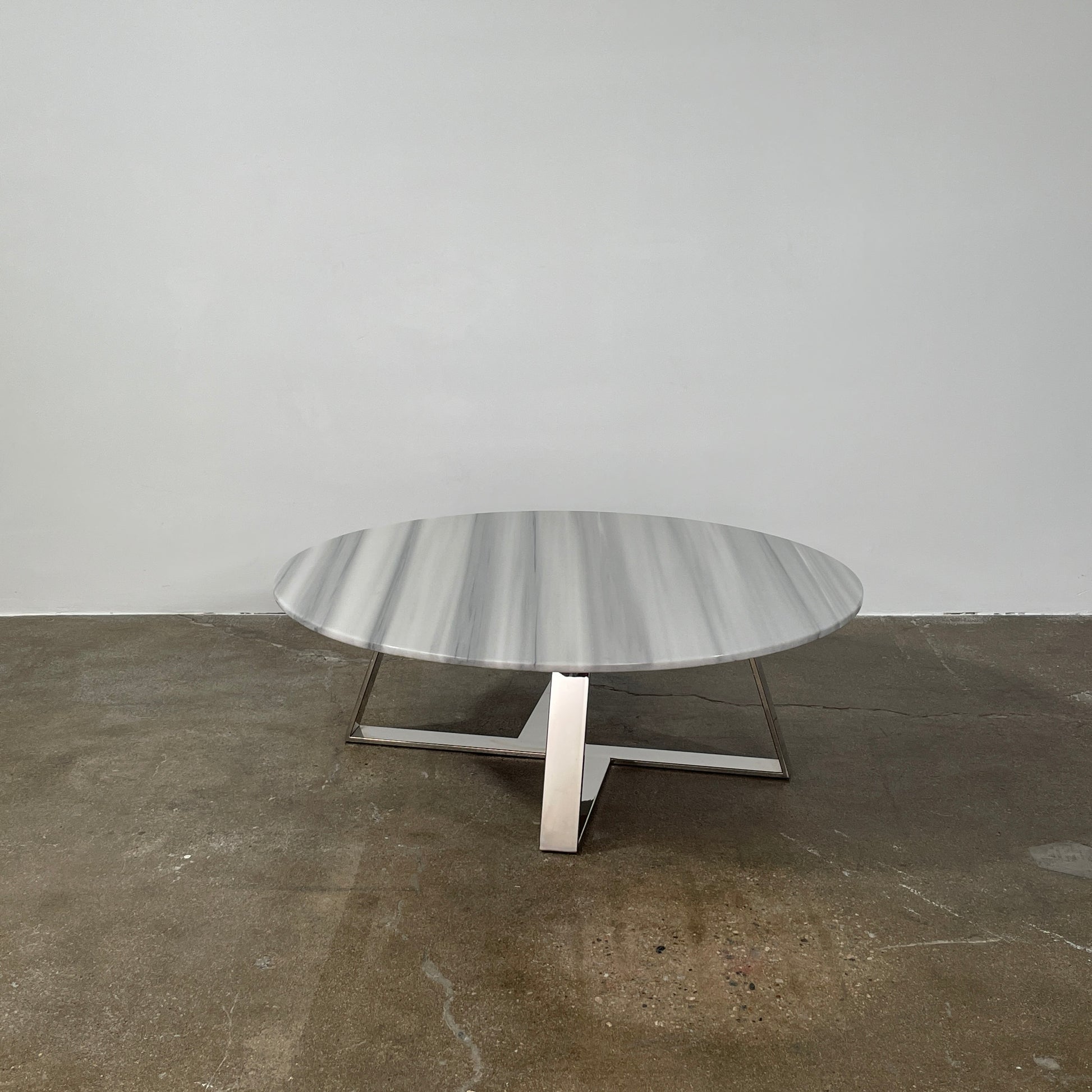 The Lievo Home Modern Studio Ingrid Coffee Table has a glossy marble top with white and gray stripes and a polished stainless steel base, shown on a bare concrete floor against a plain white wall.