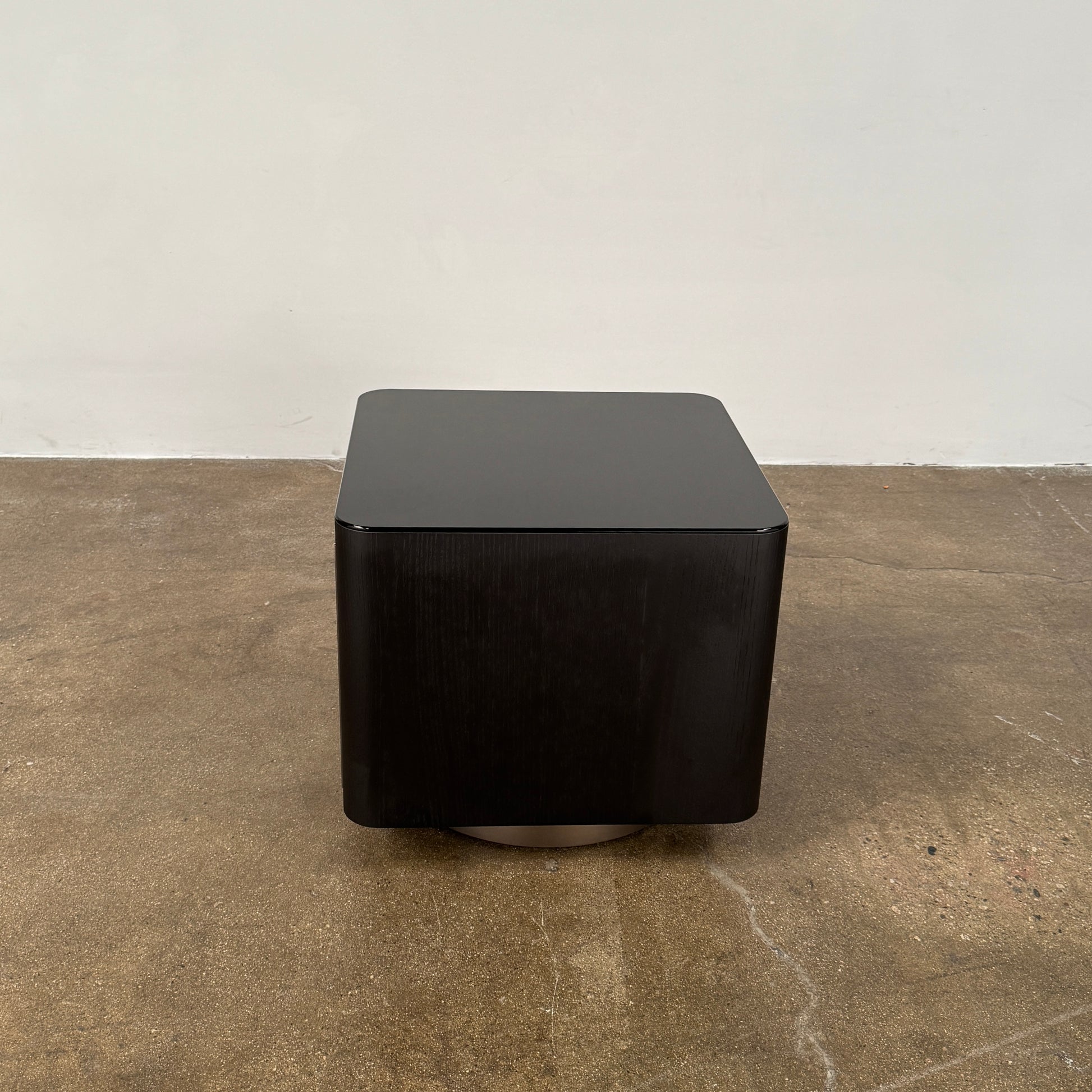 The Minotti Milton Side Table by Minotti features a modern, dark wooden cube design with an open center and a circular base, displayed on a polished concrete floor against a plain white wall.