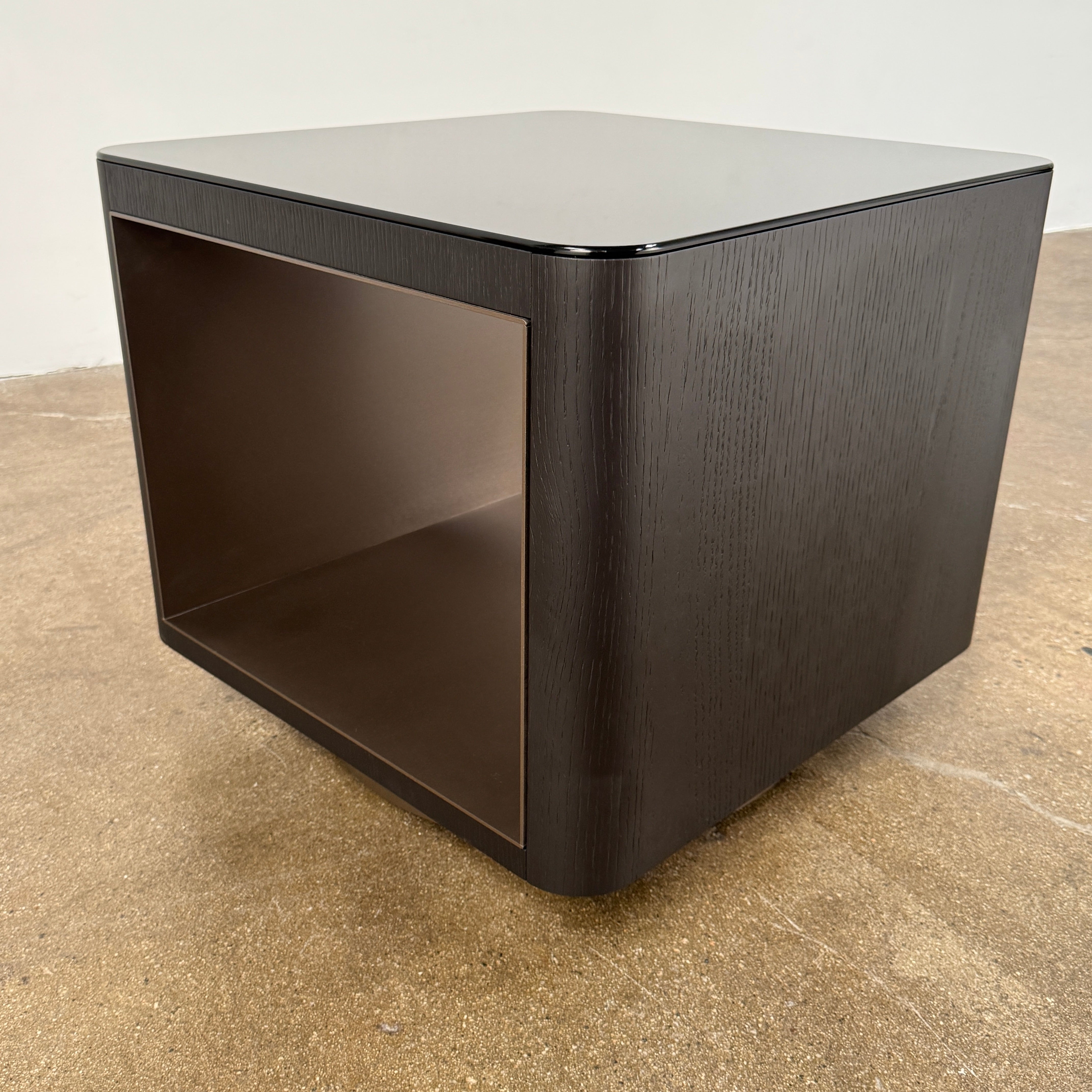 The Minotti Milton Side Table by Minotti features a modern, dark wooden cube design with an open center and a circular base, displayed on a polished concrete floor against a plain white wall.