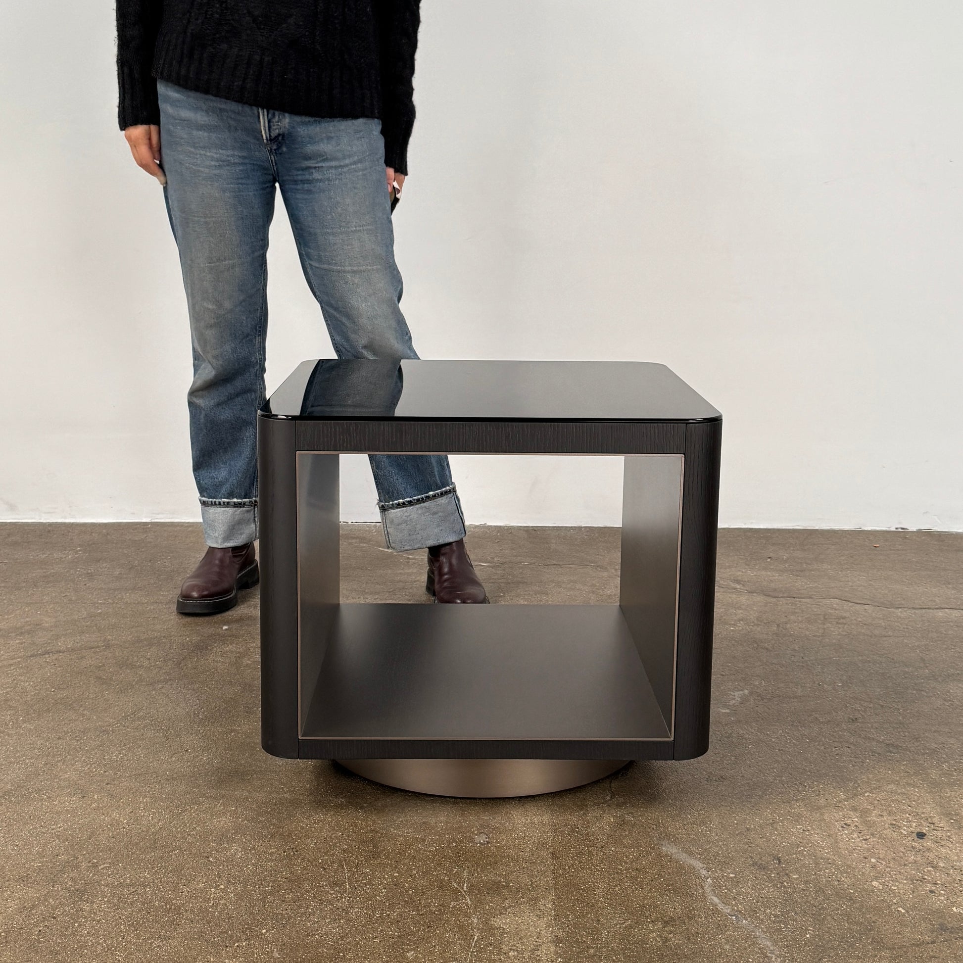 The Minotti Milton Side Table by Minotti features a modern, dark wooden cube design with an open center and a circular base, displayed on a polished concrete floor against a plain white wall.