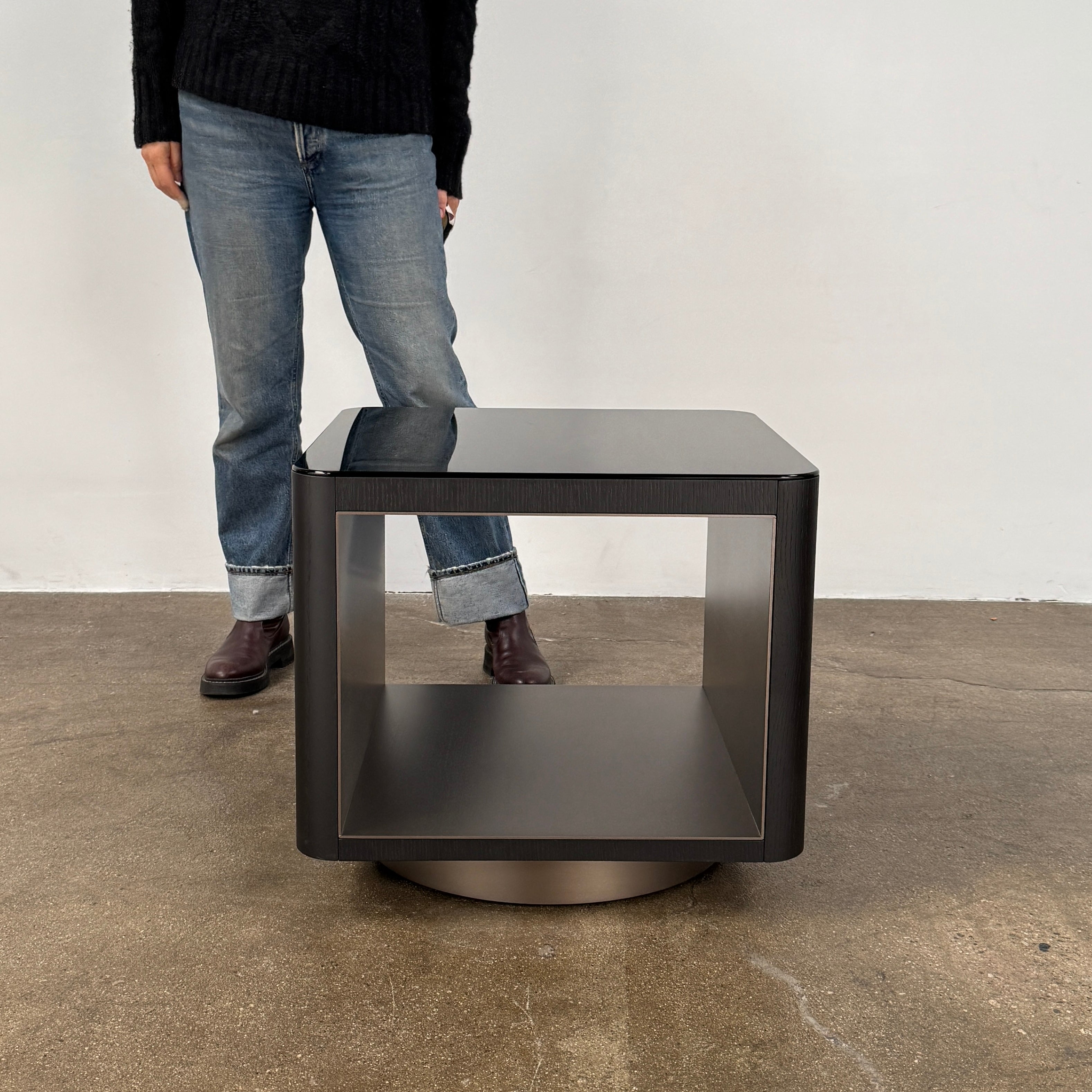 The Minotti Milton Side Table by Minotti features a modern, dark wooden cube design with an open center and a circular base, displayed on a polished concrete floor against a plain white wall.