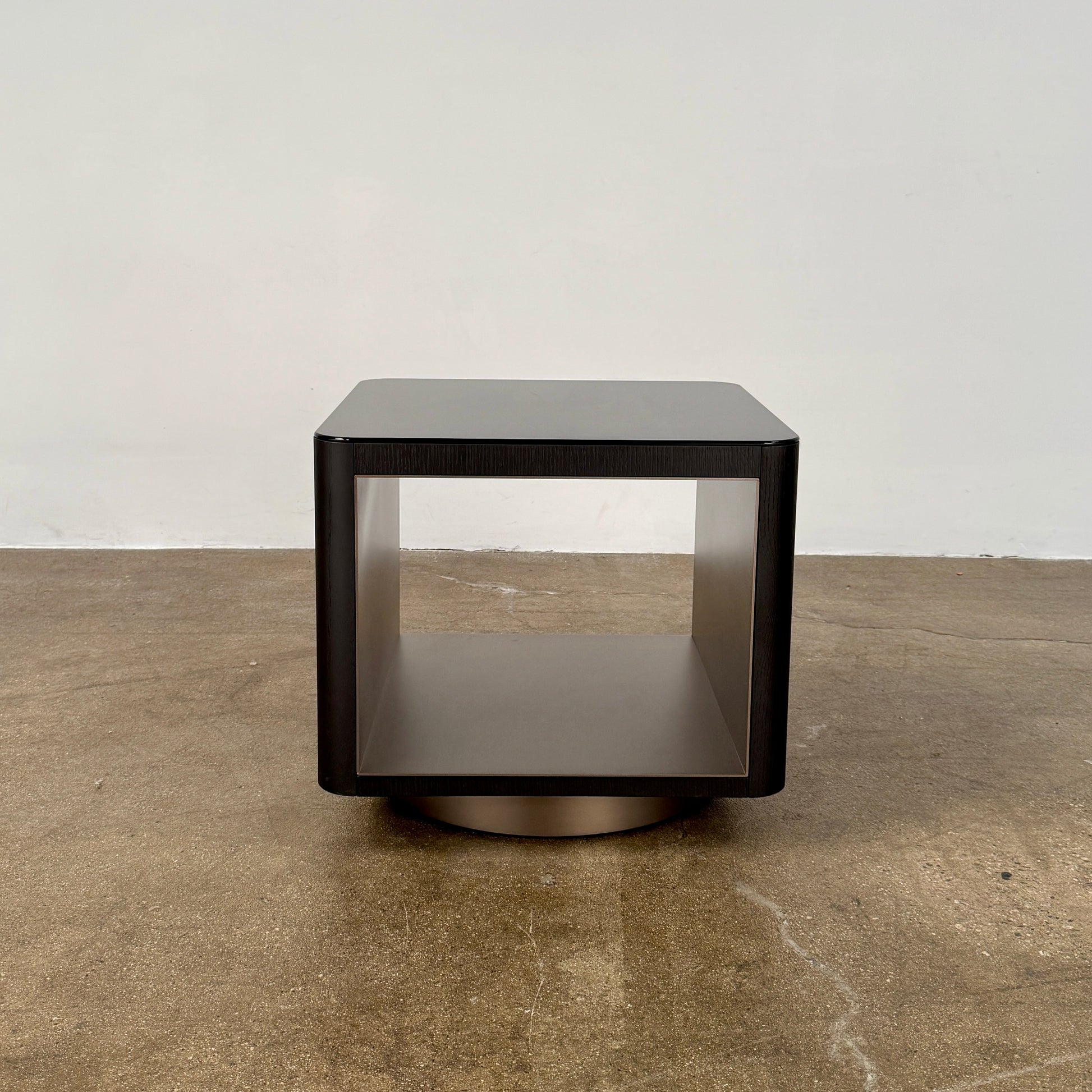 The Minotti Milton Side Table by Minotti features a modern, dark wooden cube design with an open center and a circular base, displayed on a polished concrete floor against a plain white wall.