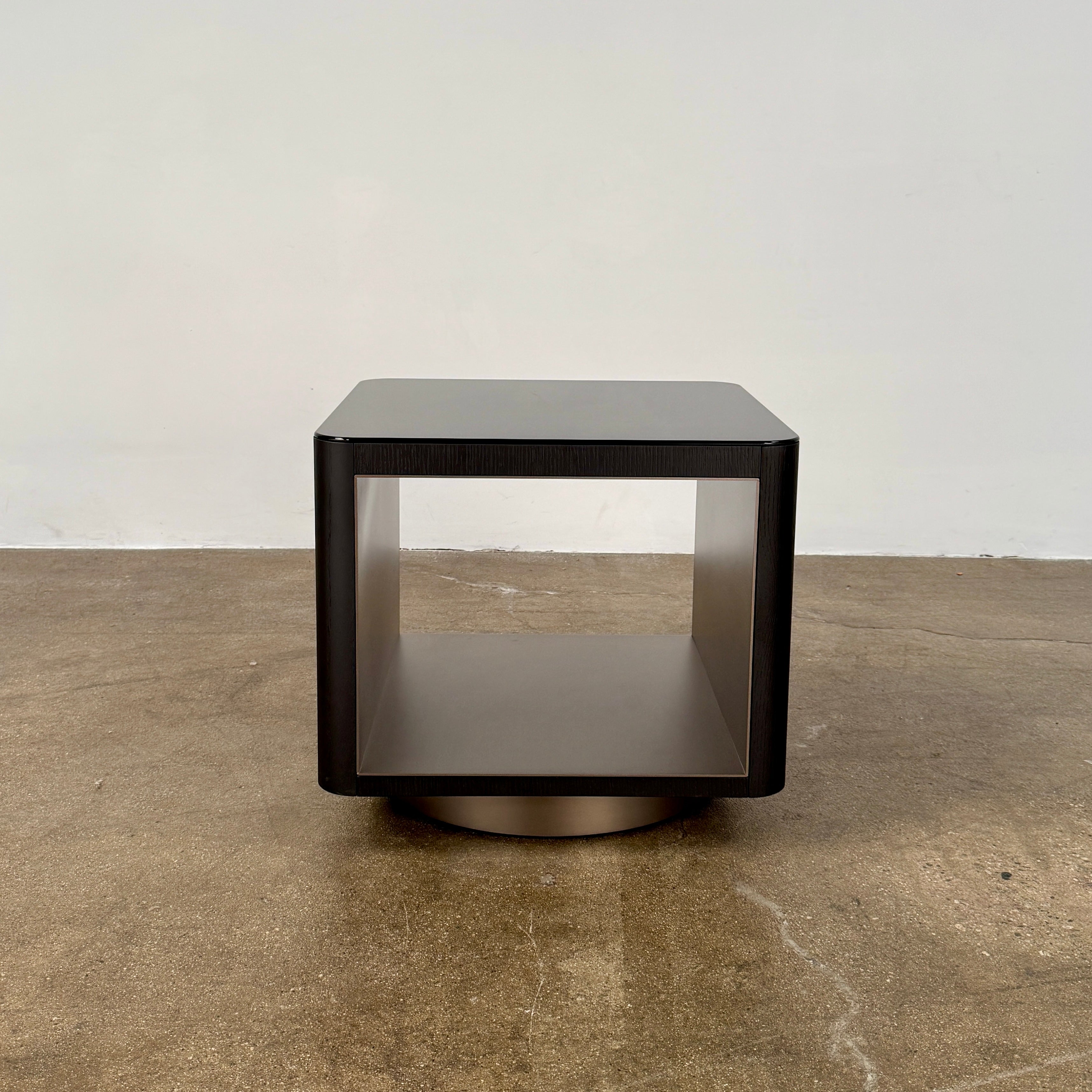The Minotti Milton Side Table by Minotti features a modern, dark wooden cube design with an open center and a circular base, displayed on a polished concrete floor against a plain white wall.