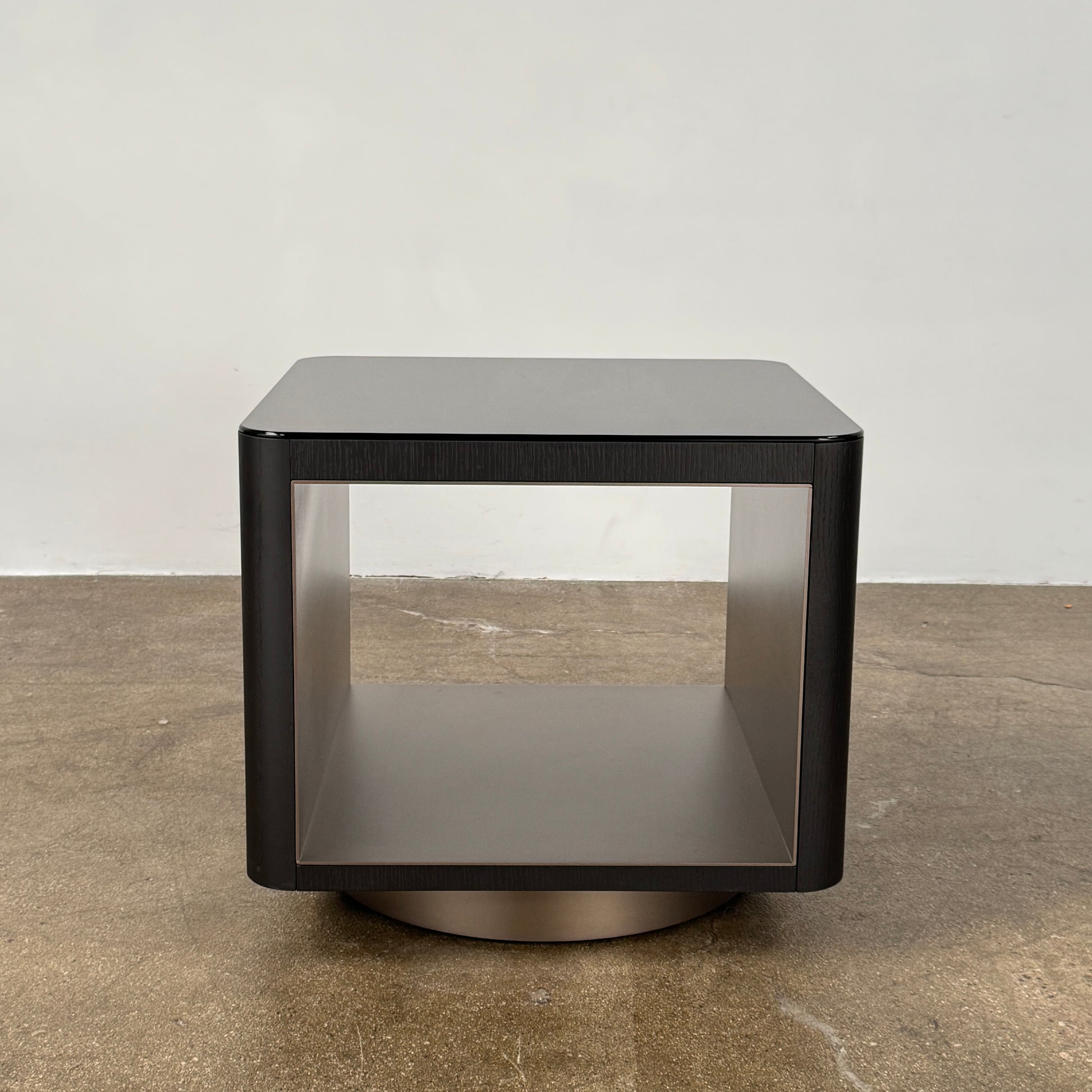 The Minotti Milton Side Table by Minotti features a modern, dark wooden cube design with an open center and a circular base, displayed on a polished concrete floor against a plain white wall.