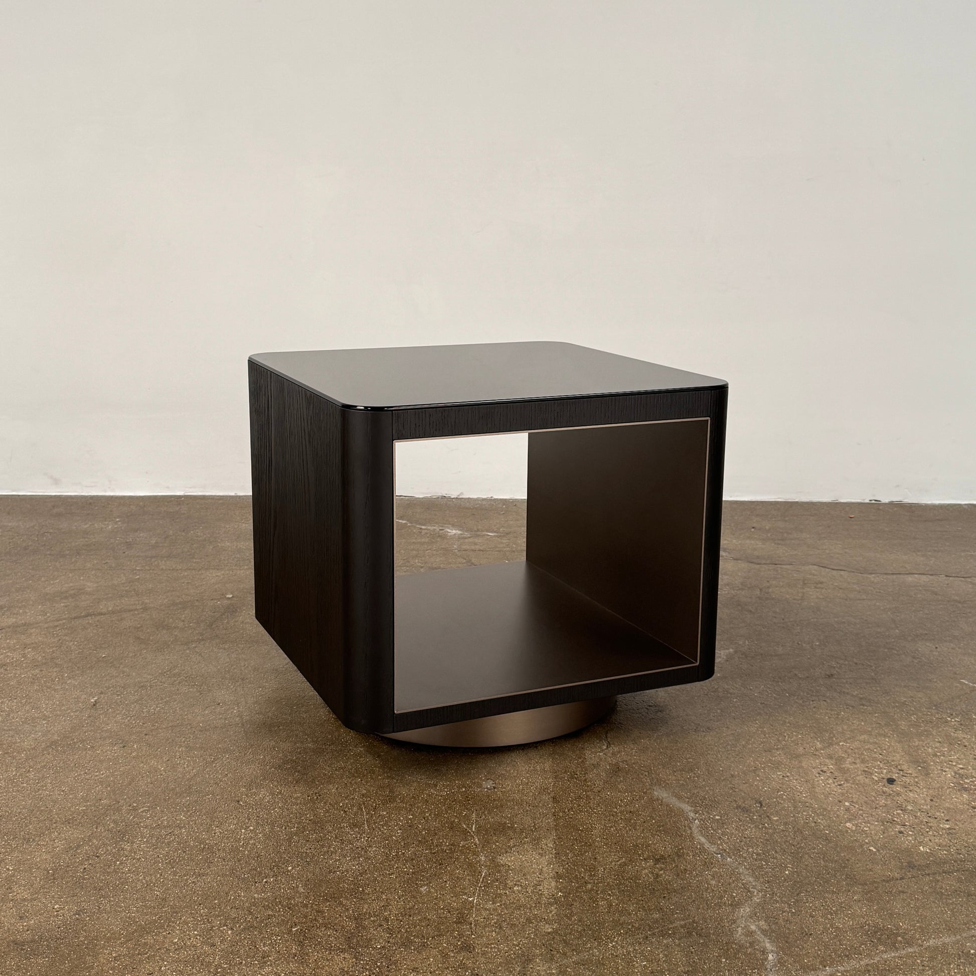 The Minotti Milton Side Table by Minotti features a modern, dark wooden cube design with an open center and a circular base, displayed on a polished concrete floor against a plain white wall.