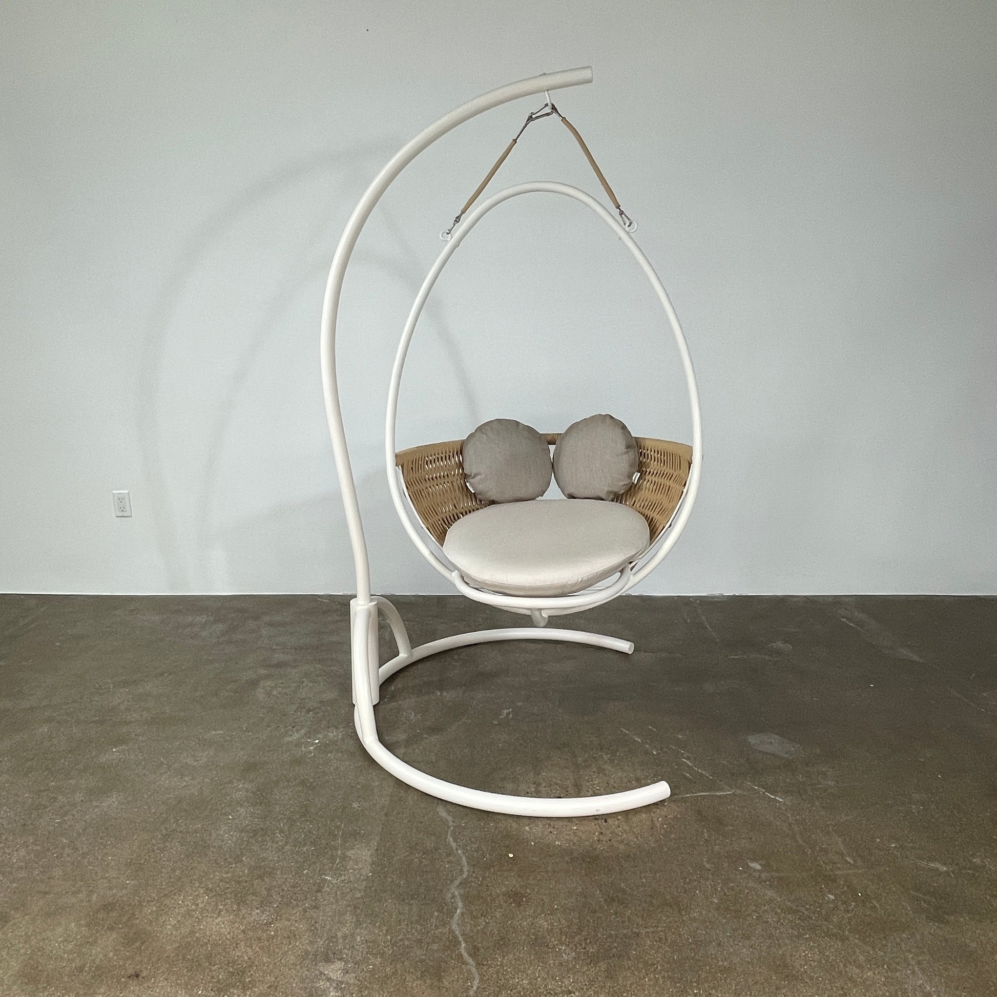 The Point 1920 Weave Hanging Swing with Base by Modern Resale features a white metal frame, wicker seat, and beige cushions, shown freestanding and isolated on a plain white background.