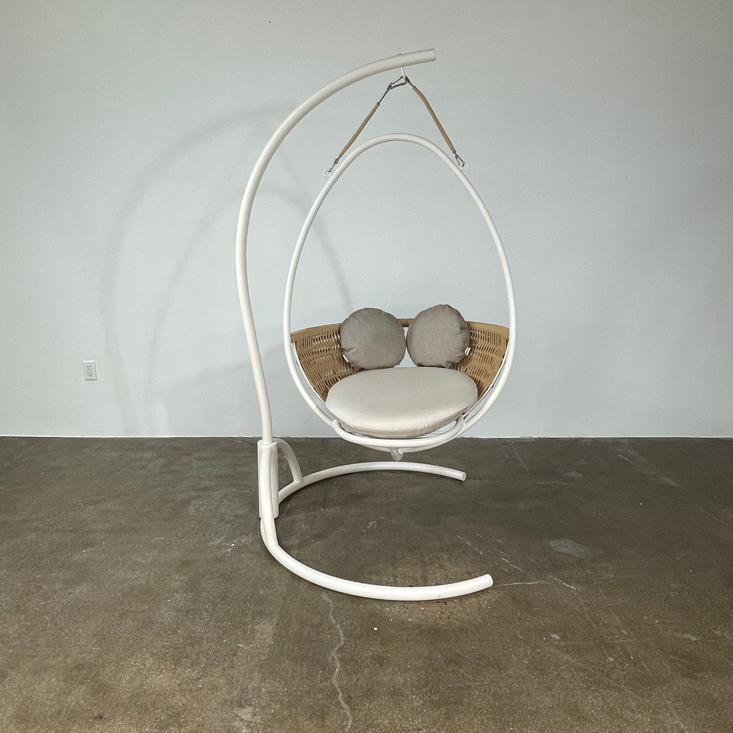 The Point 1920 Weave Hanging Swing with Base by Modern Resale features a white metal frame, wicker seat, and beige cushions, shown freestanding and isolated on a plain white background.