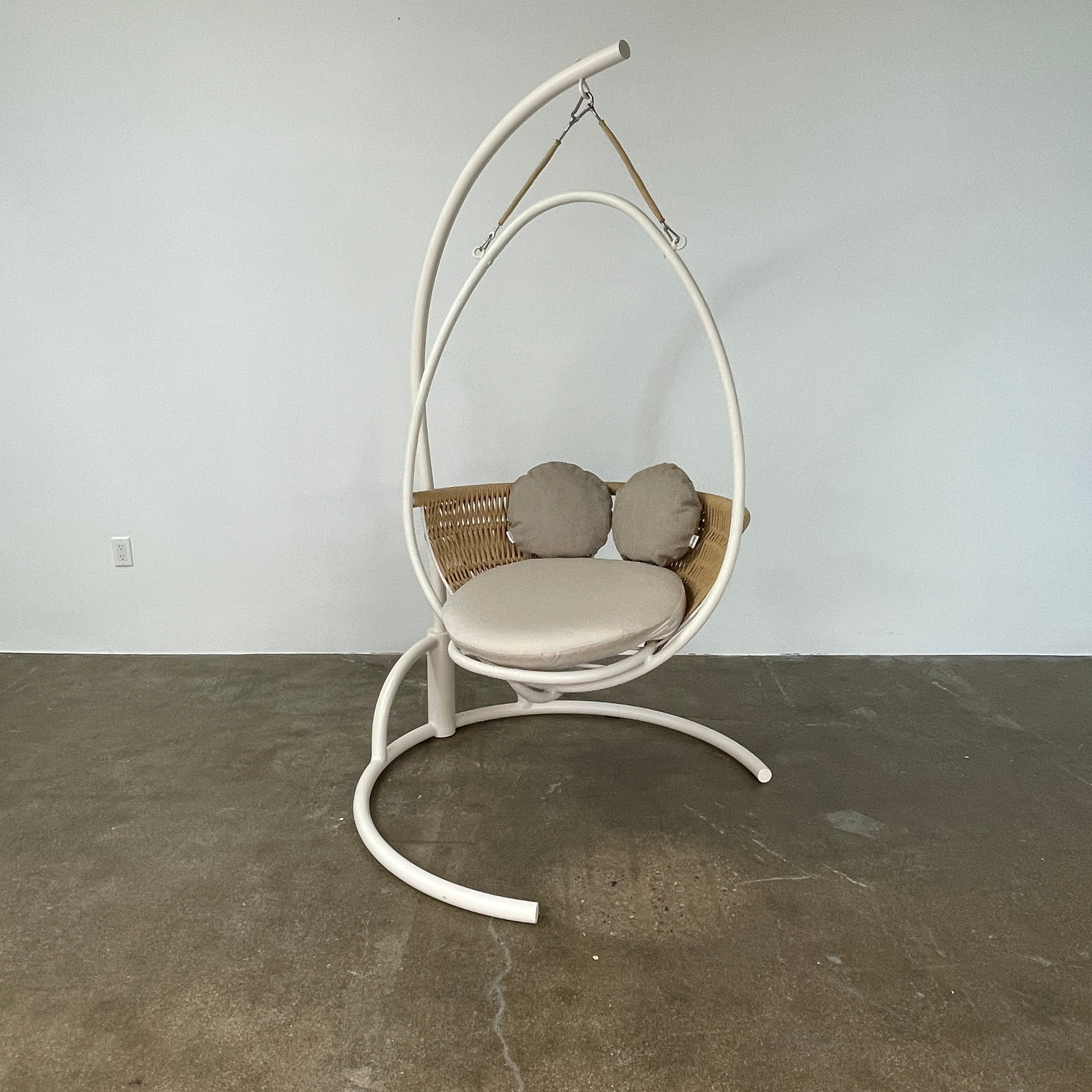 The Point 1920 Weave Hanging Swing with Base by Modern Resale features a white metal frame, wicker seat, and beige cushions, shown freestanding and isolated on a plain white background.