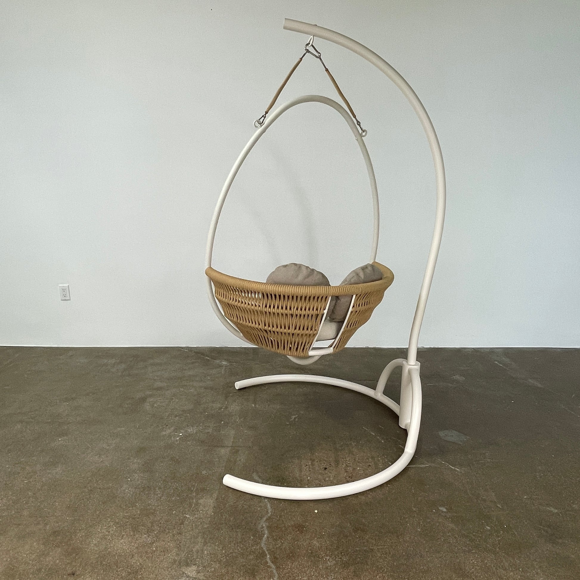 The Point 1920 Weave Hanging Swing with Base by Modern Resale features a white metal frame, wicker seat, and beige cushions, shown freestanding and isolated on a plain white background.