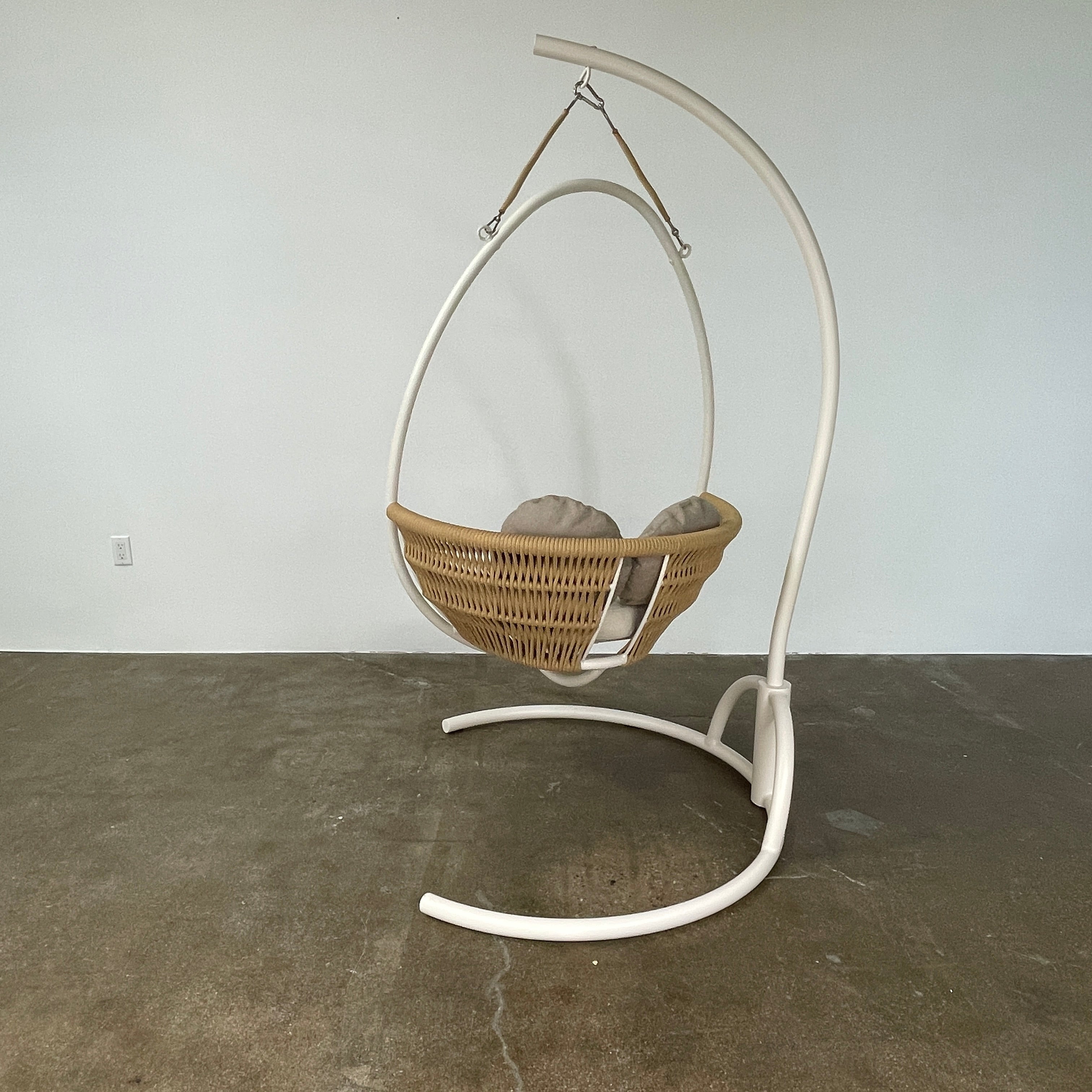 The Point 1920 Weave Hanging Swing with Base by Modern Resale features a white metal frame, wicker seat, and beige cushions, shown freestanding and isolated on a plain white background.
