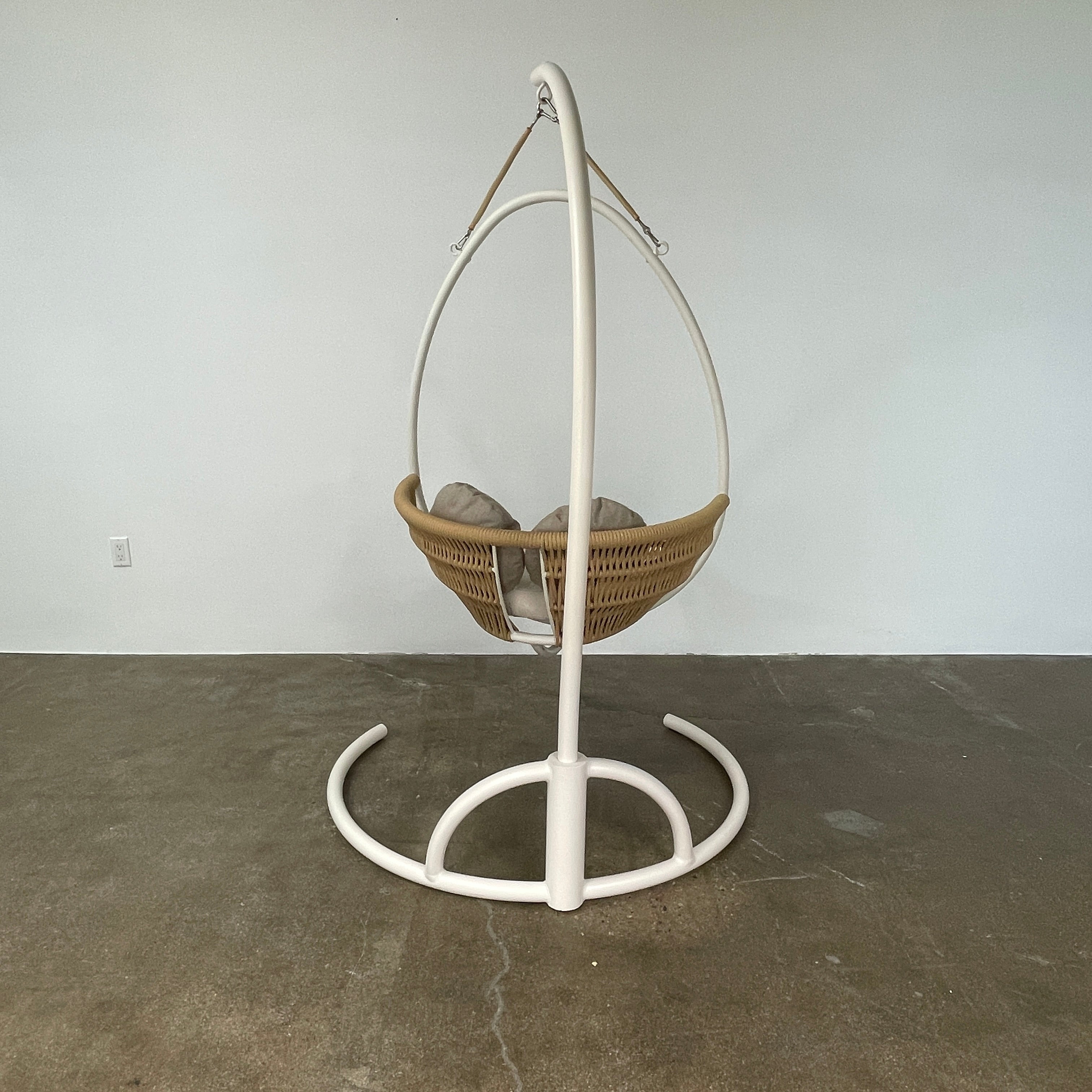 The Point 1920 Weave Hanging Swing with Base by Modern Resale features a white metal frame, wicker seat, and beige cushions, shown freestanding and isolated on a plain white background.