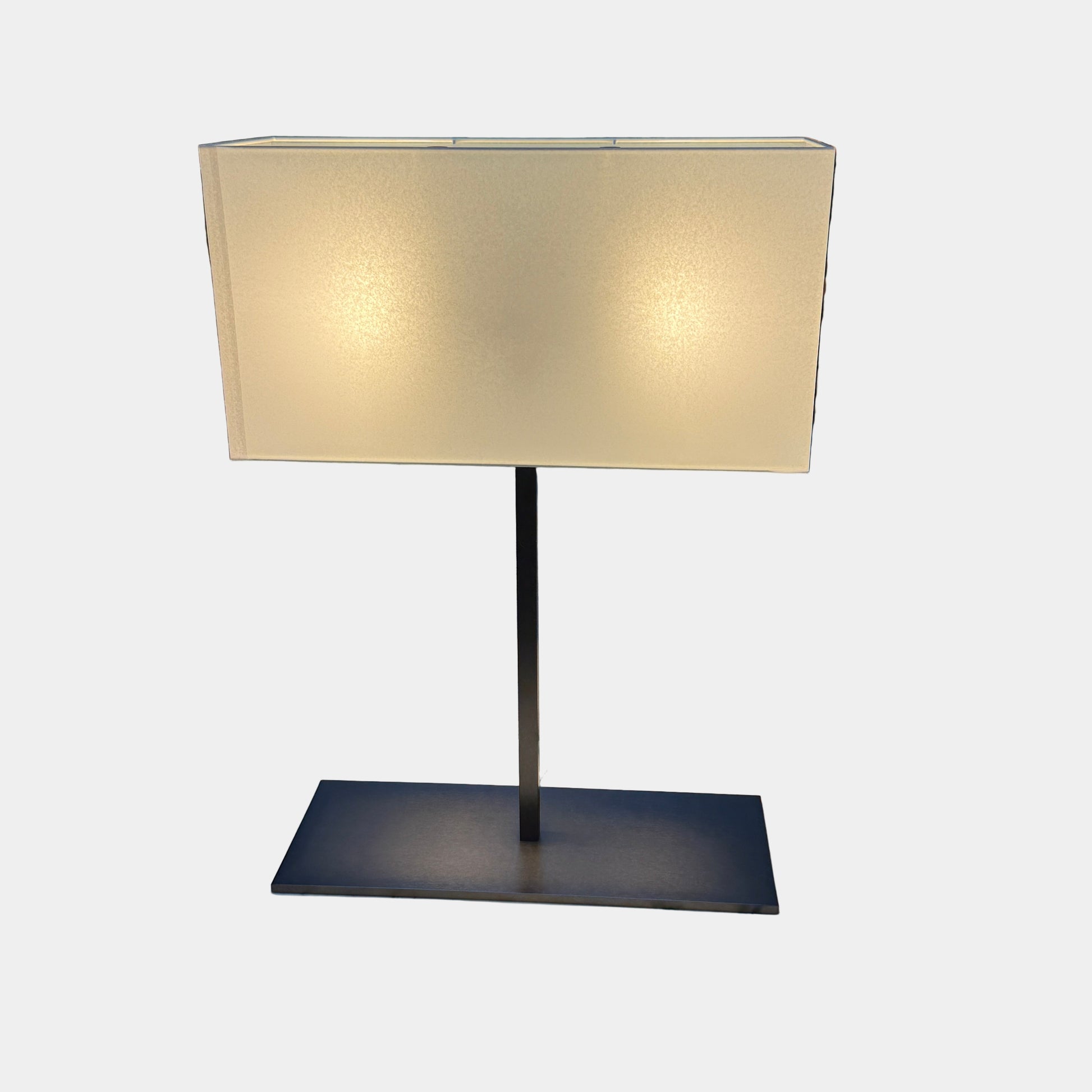The Maxalto Leukon Table Lamp by Maxalto features a rectangular fabric shade, slim metal stem, and flat base, providing warm, modern lighting.