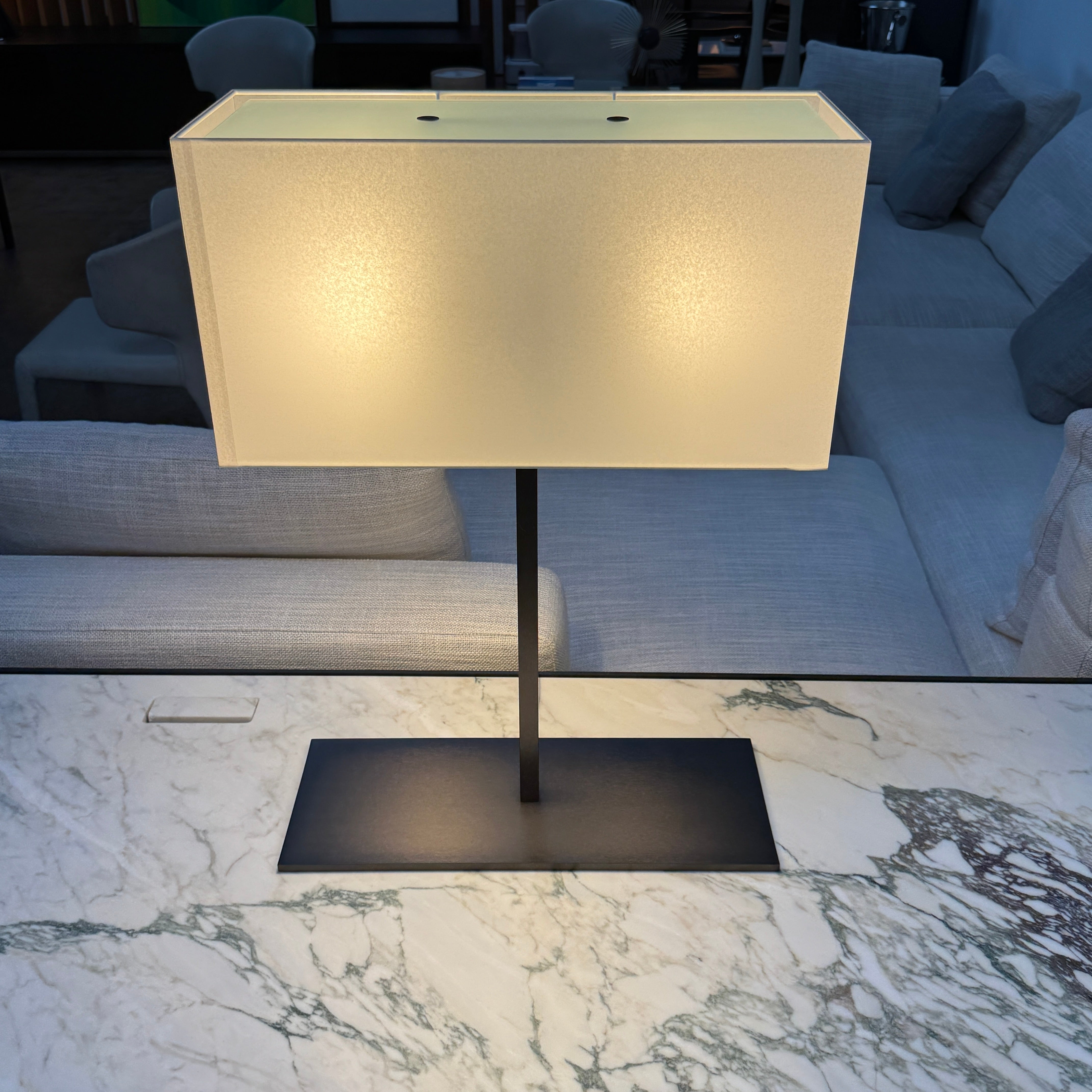 The Maxalto Leukon Table Lamp by Maxalto features a rectangular fabric shade, slim metal stem, and flat base, providing warm, modern lighting.