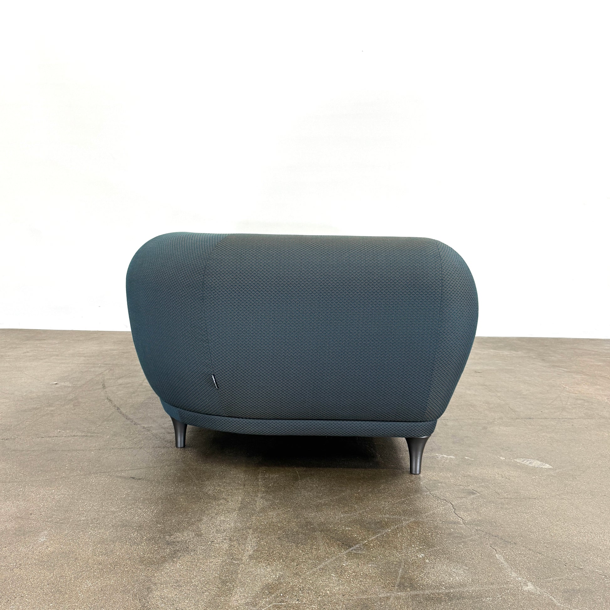 A Roche Bobois Montgolfiere sofa in dark gray, featuring rounded arms and black legs, from the Globe Trotter collection by Marcel Wanders, sits on a polished concrete floor against a plain white wall.