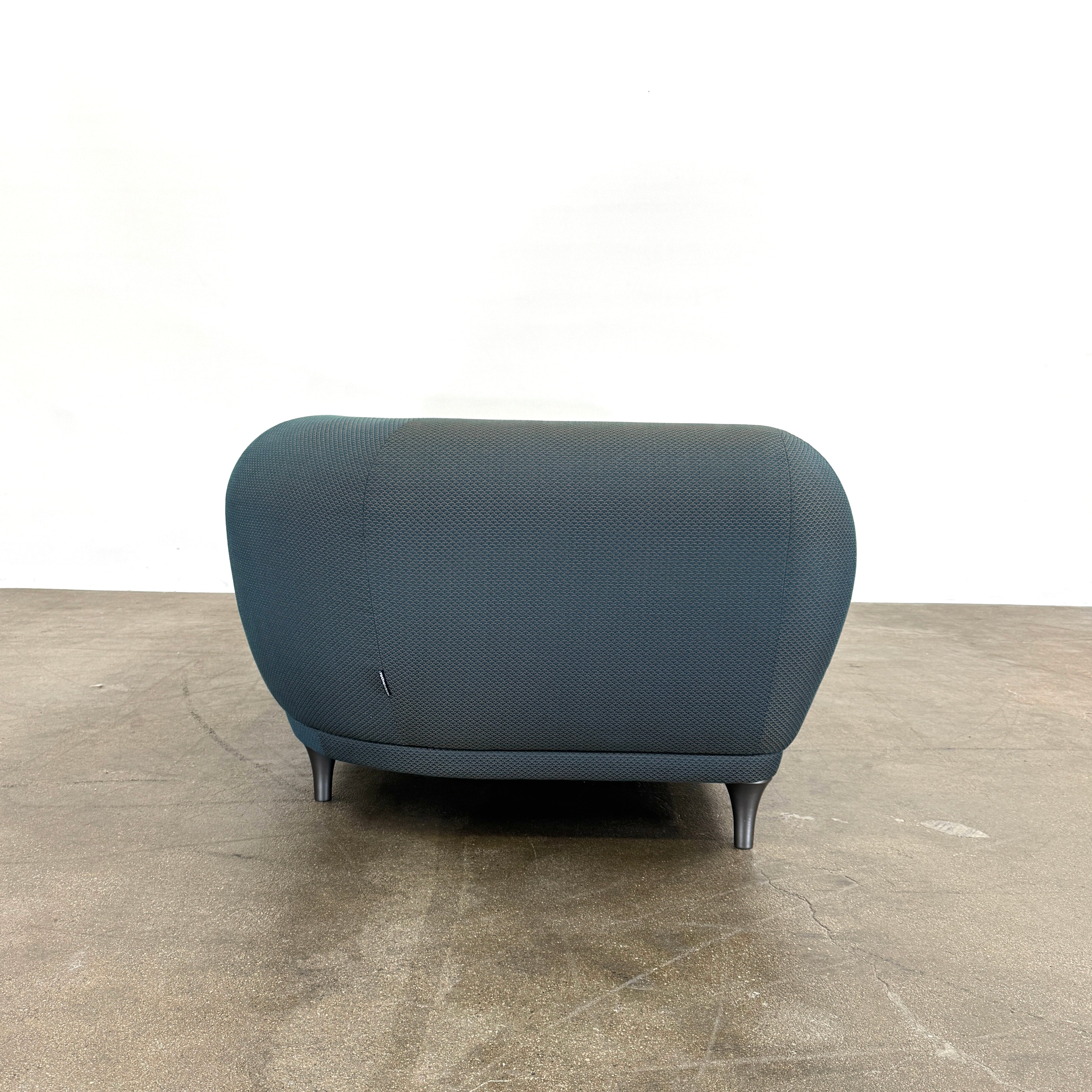 A Roche Bobois Montgolfiere sofa in dark gray, featuring rounded arms and black legs, from the Globe Trotter collection by Marcel Wanders, sits on a polished concrete floor against a plain white wall.