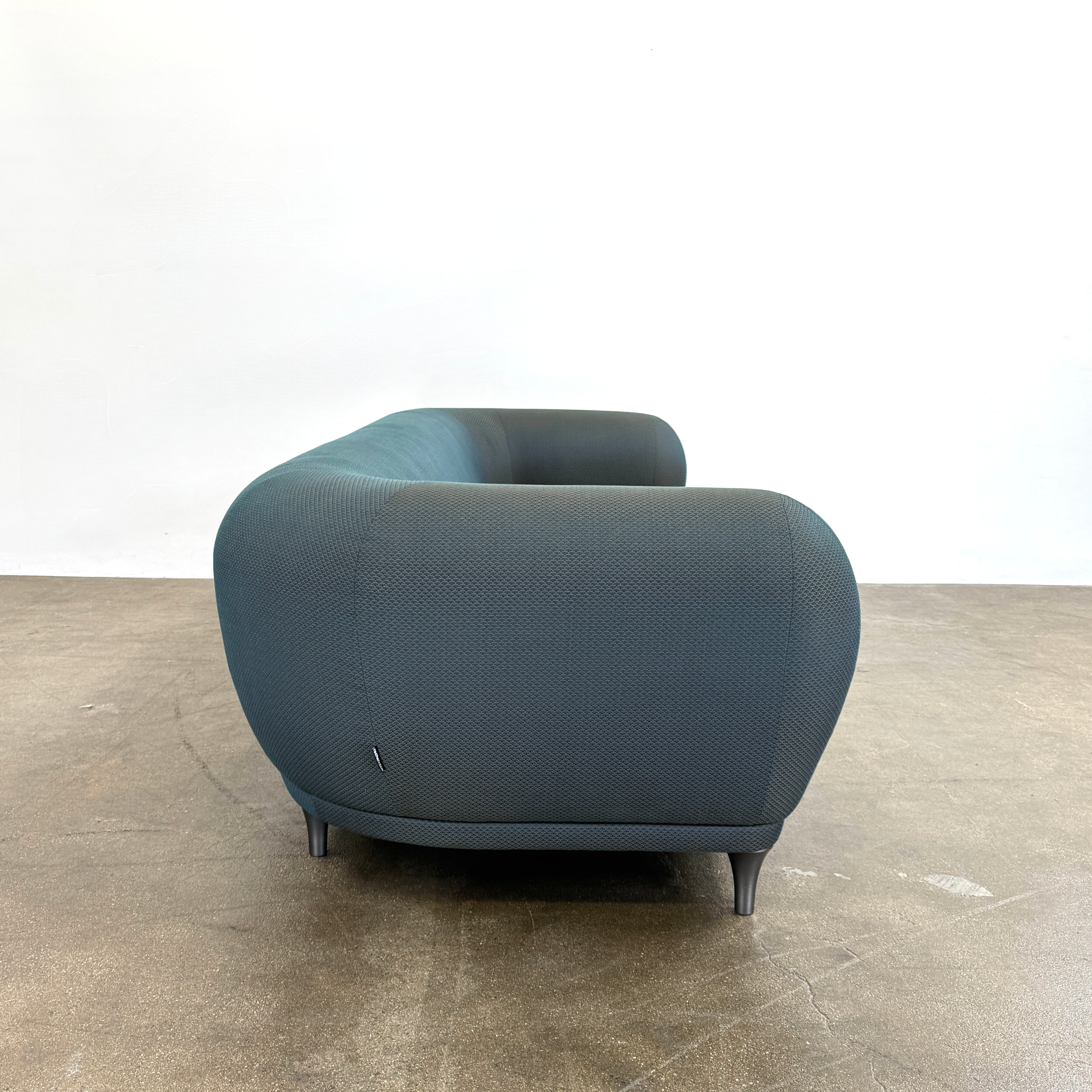 A Roche Bobois Montgolfiere sofa in dark gray, featuring rounded arms and black legs, from the Globe Trotter collection by Marcel Wanders, sits on a polished concrete floor against a plain white wall.