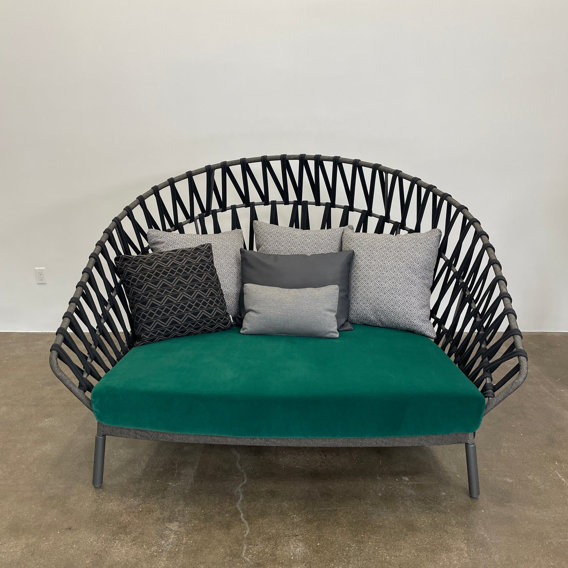A Varaschin Outdoor Emma Cross Daybed in black with a green cushion and five assorted gray and black pillows rests on a concrete floor by a plain white wall.