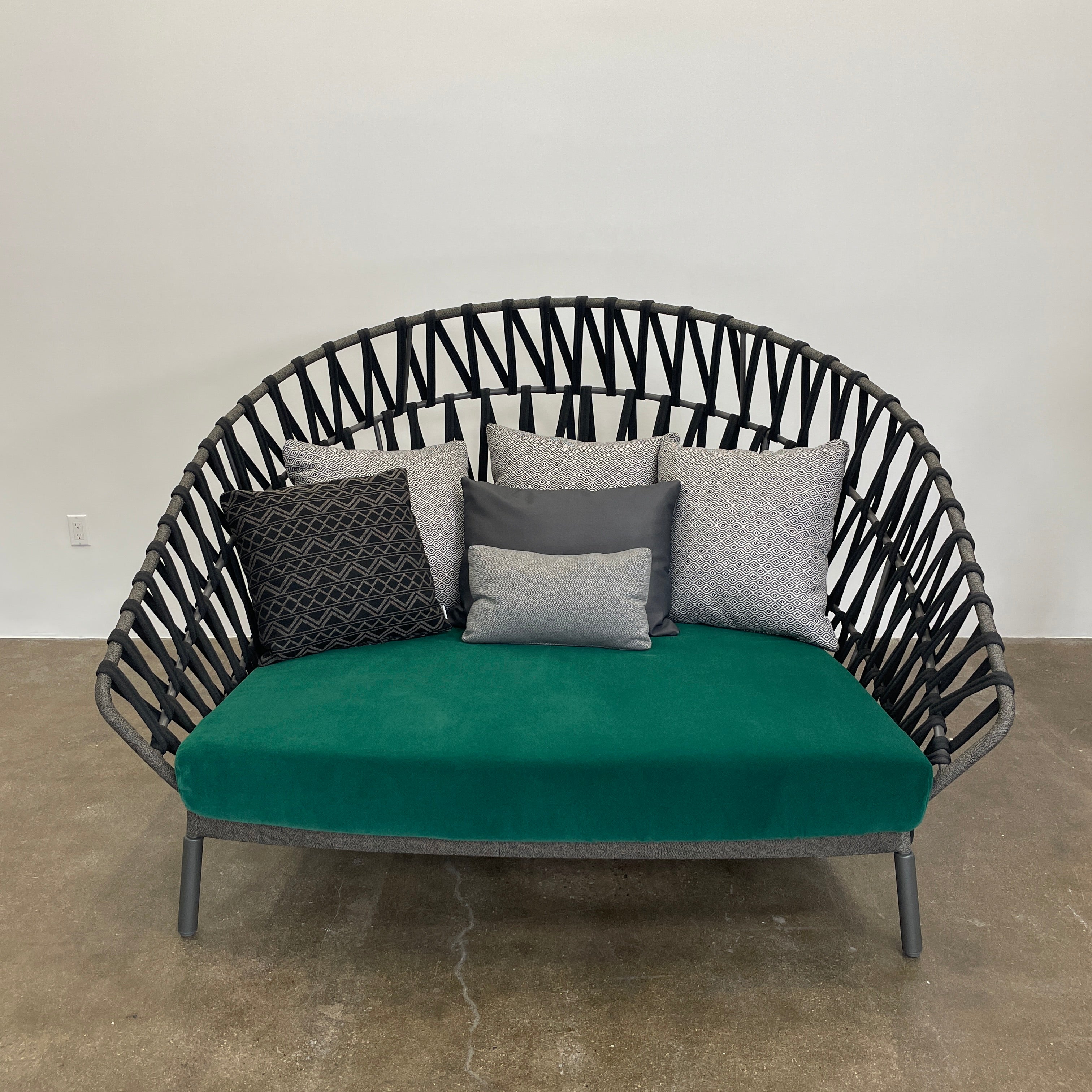 A Varaschin Outdoor Emma Cross Daybed in black with a green cushion and five assorted gray and black pillows rests on a concrete floor by a plain white wall.