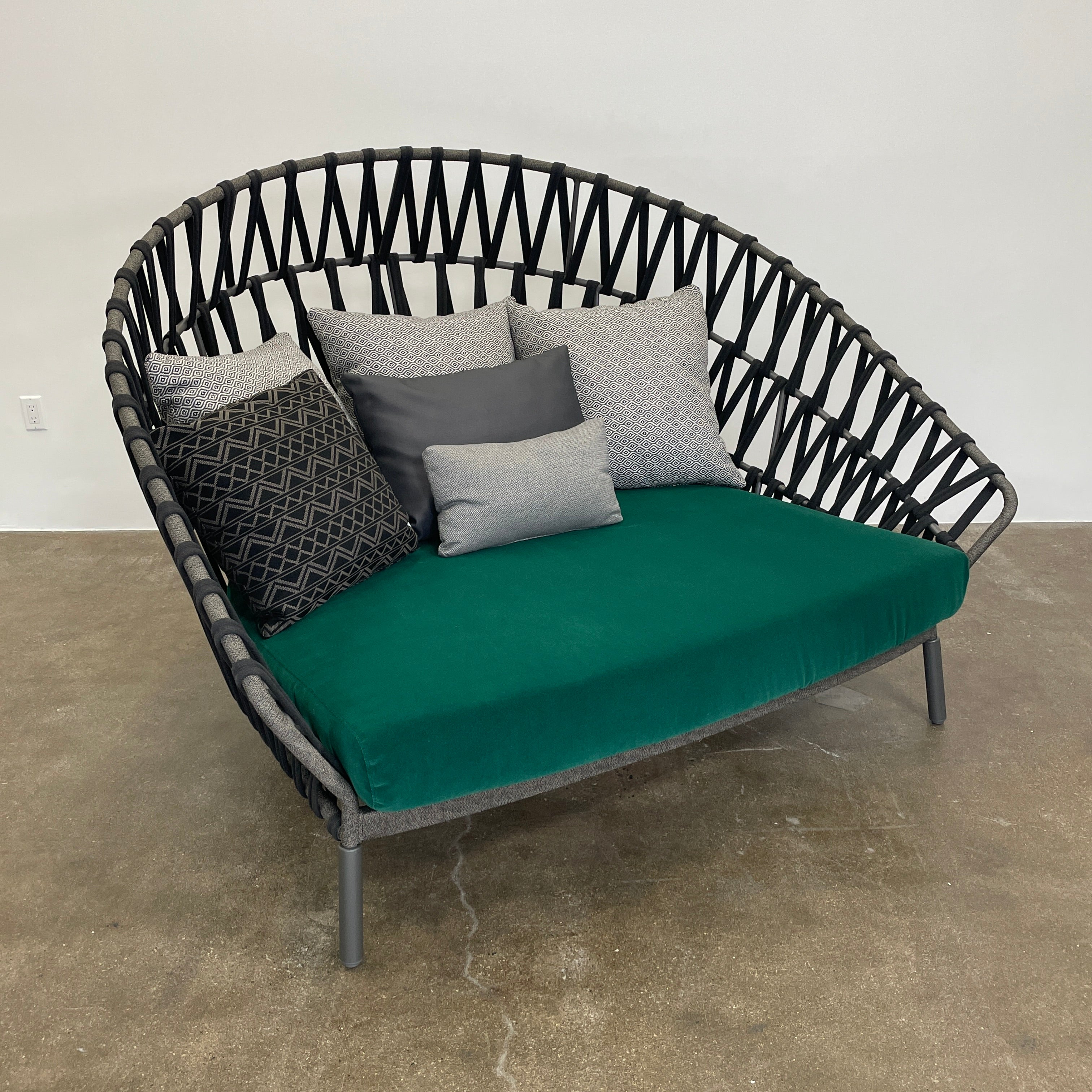 A Varaschin Outdoor Emma Cross Daybed in black with a green cushion and five assorted gray and black pillows rests on a concrete floor by a plain white wall.