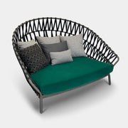 A Varaschin Outdoor Emma Cross Daybed in black with a green cushion and five assorted gray and black pillows rests on a concrete floor by a plain white wall.