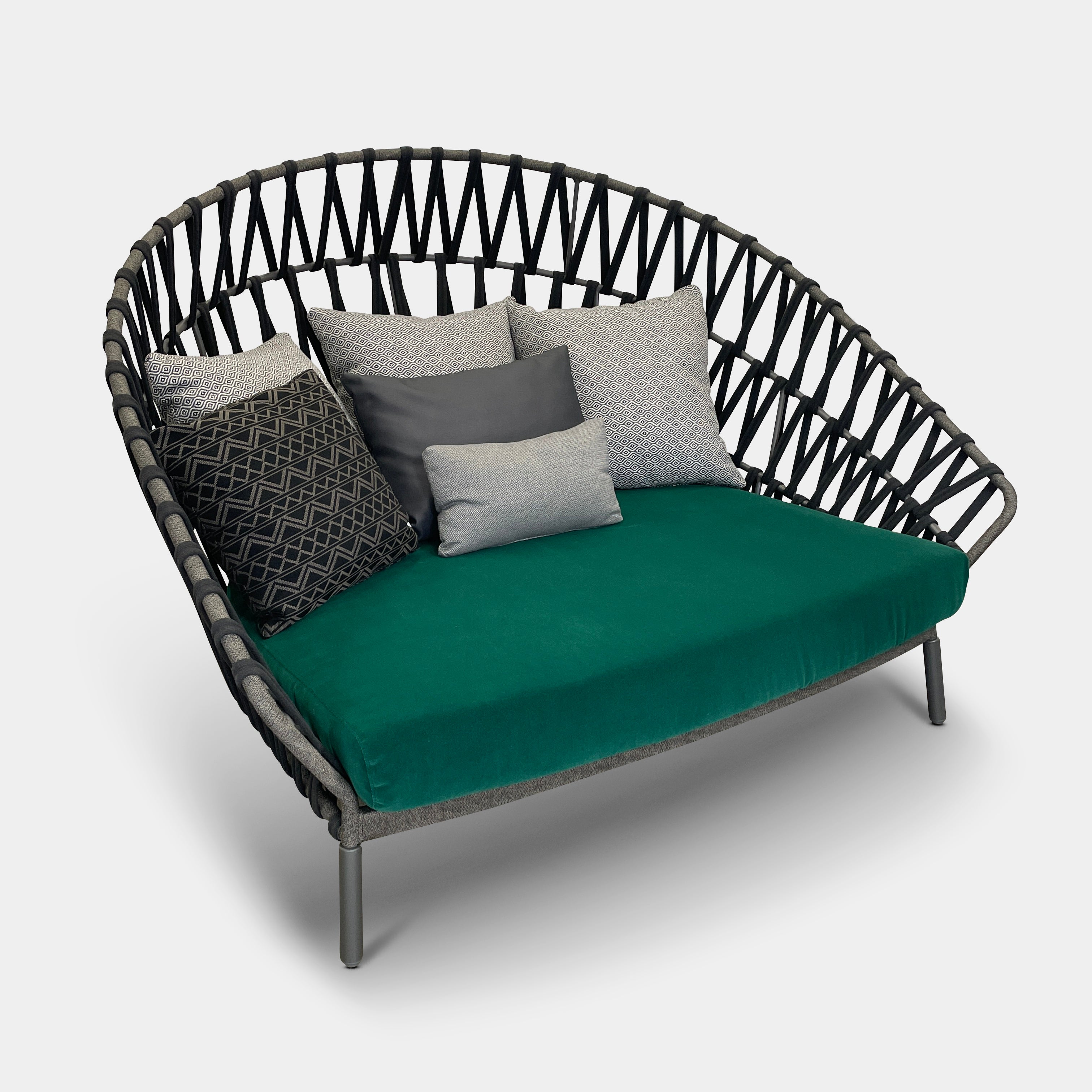 A Varaschin Outdoor Emma Cross Daybed in black with a green cushion and five assorted gray and black pillows rests on a concrete floor by a plain white wall.
