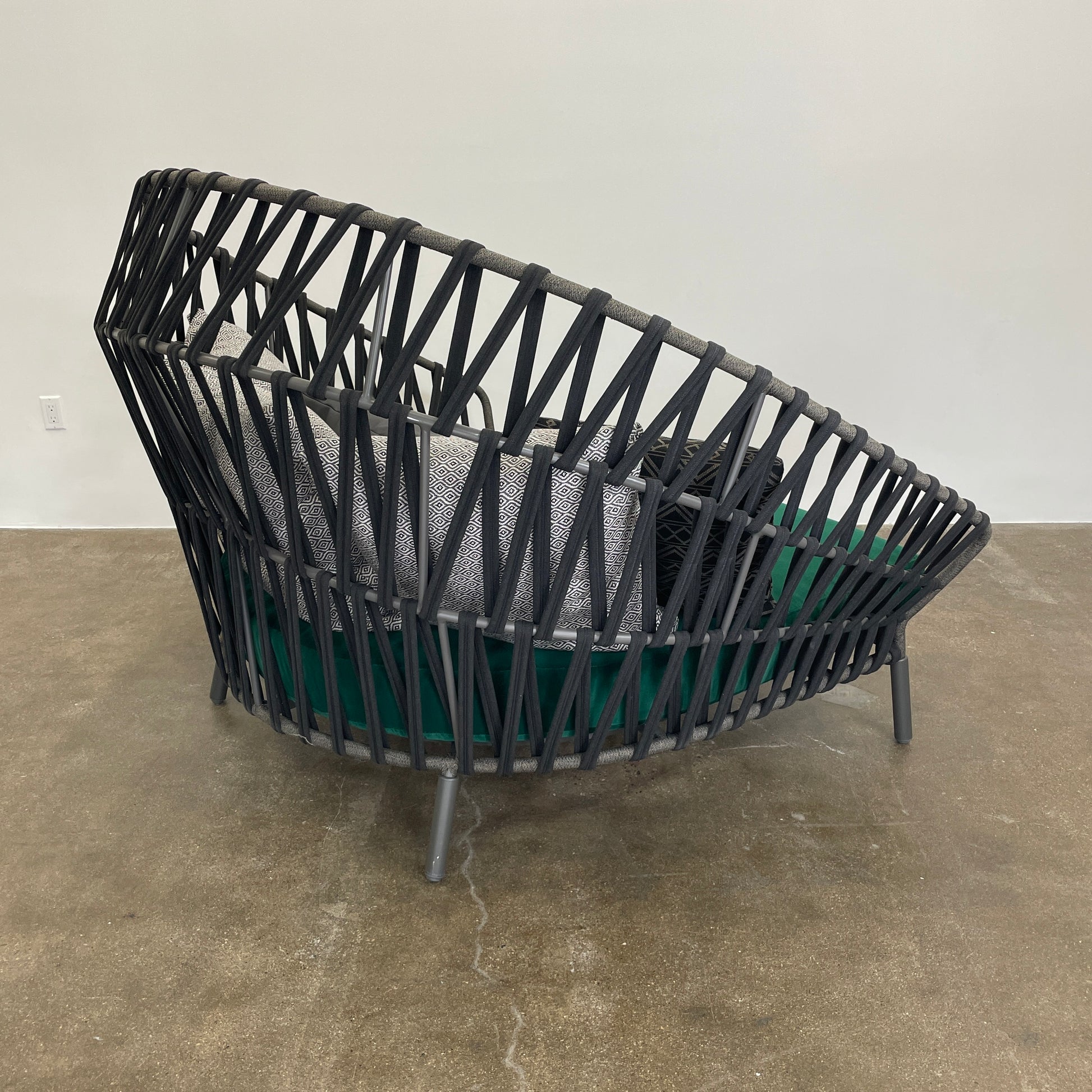 A Varaschin Outdoor Emma Cross Daybed in black with a green cushion and five assorted gray and black pillows rests on a concrete floor by a plain white wall.