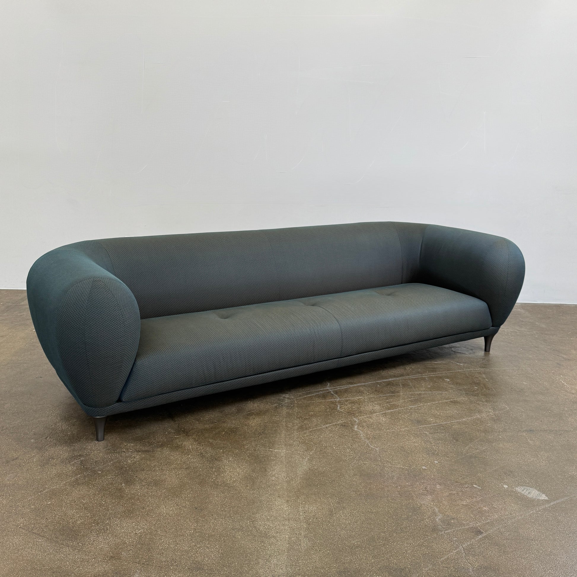 A Roche Bobois Montgolfiere sofa in dark gray, featuring rounded arms and black legs, from the Globe Trotter collection by Marcel Wanders, sits on a polished concrete floor against a plain white wall.