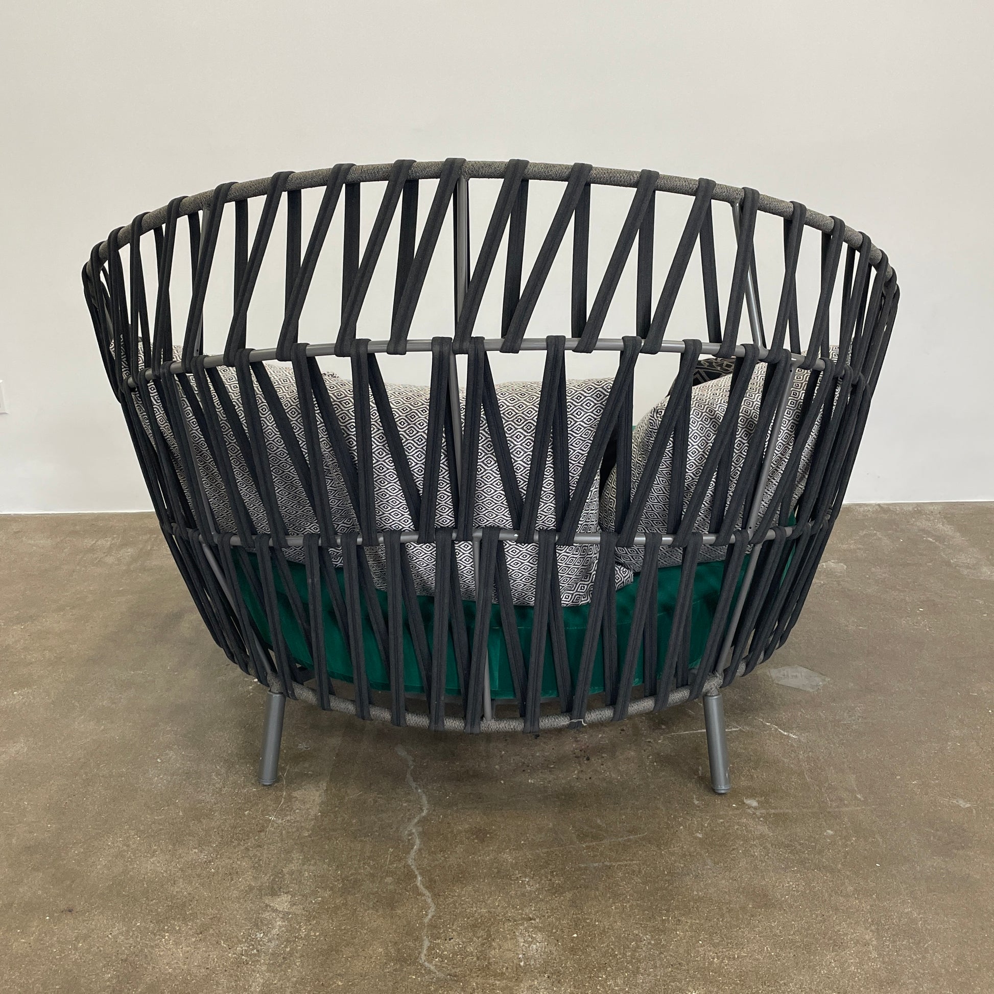 A Varaschin Outdoor Emma Cross Daybed in black with a green cushion and five assorted gray and black pillows rests on a concrete floor by a plain white wall.