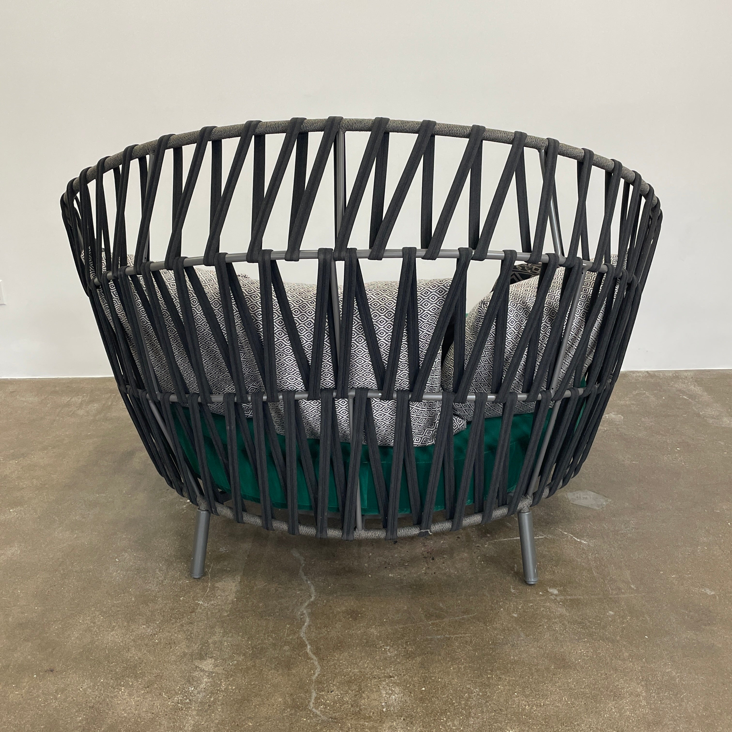 A Varaschin Outdoor Emma Cross Daybed in black with a green cushion and five assorted gray and black pillows rests on a concrete floor by a plain white wall.