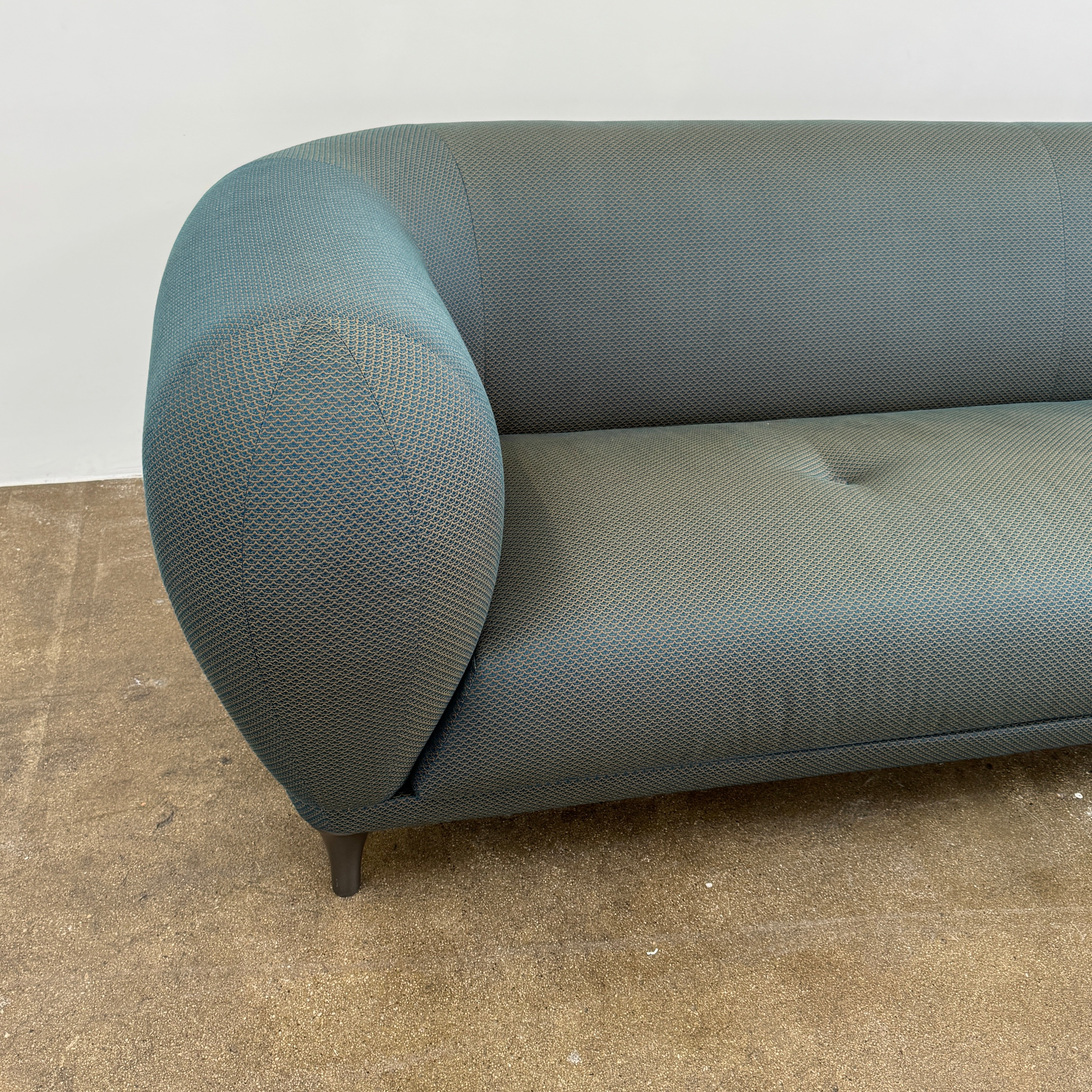 A Roche Bobois Montgolfiere sofa in dark gray, featuring rounded arms and black legs, from the Globe Trotter collection by Marcel Wanders, sits on a polished concrete floor against a plain white wall.