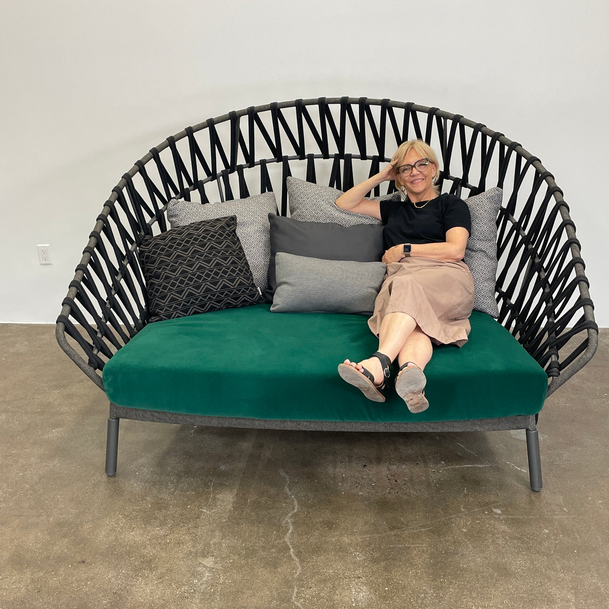A Varaschin Outdoor Emma Cross Daybed in black with a green cushion and five assorted gray and black pillows rests on a concrete floor by a plain white wall.