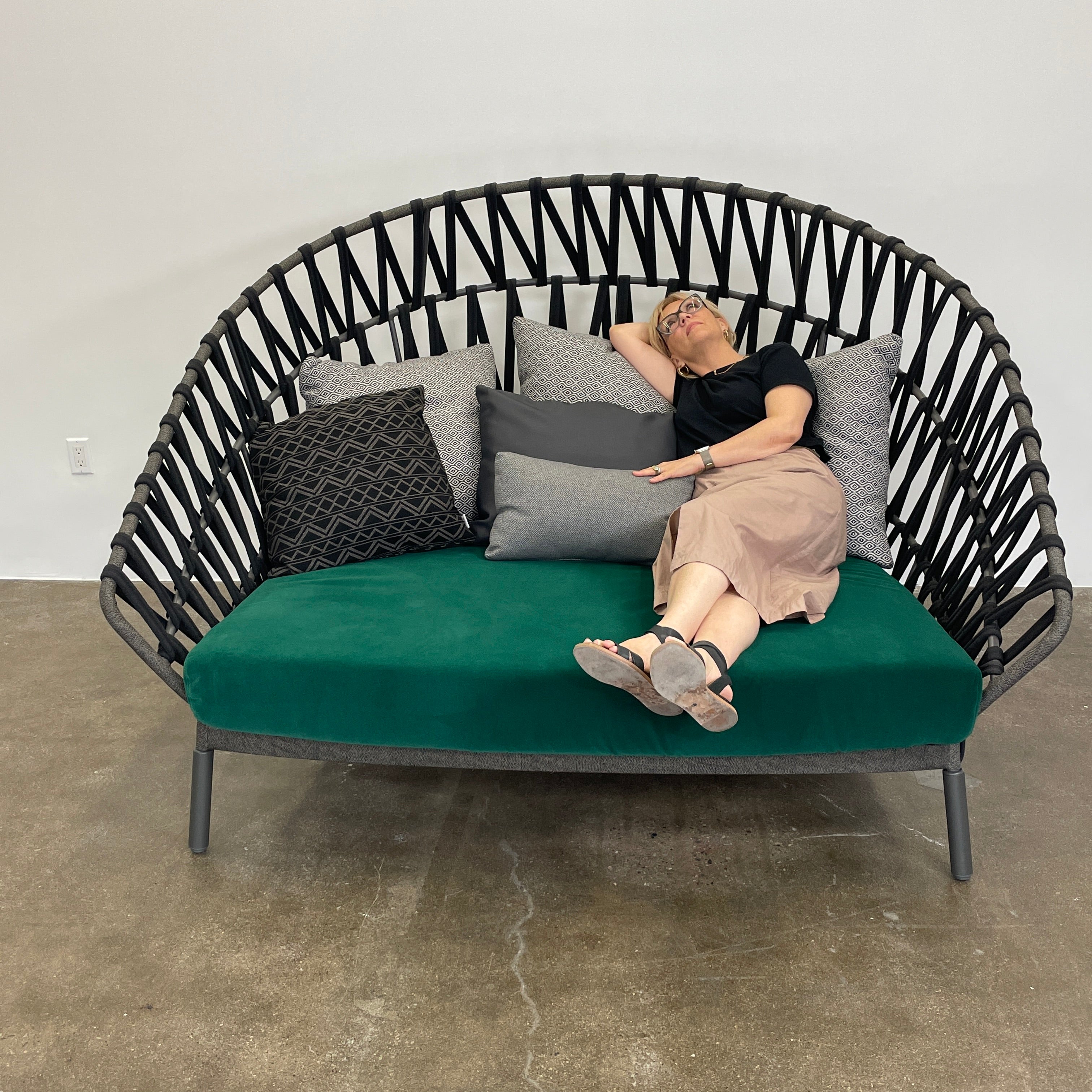 A Varaschin Outdoor Emma Cross Daybed in black with a green cushion and five assorted gray and black pillows rests on a concrete floor by a plain white wall.