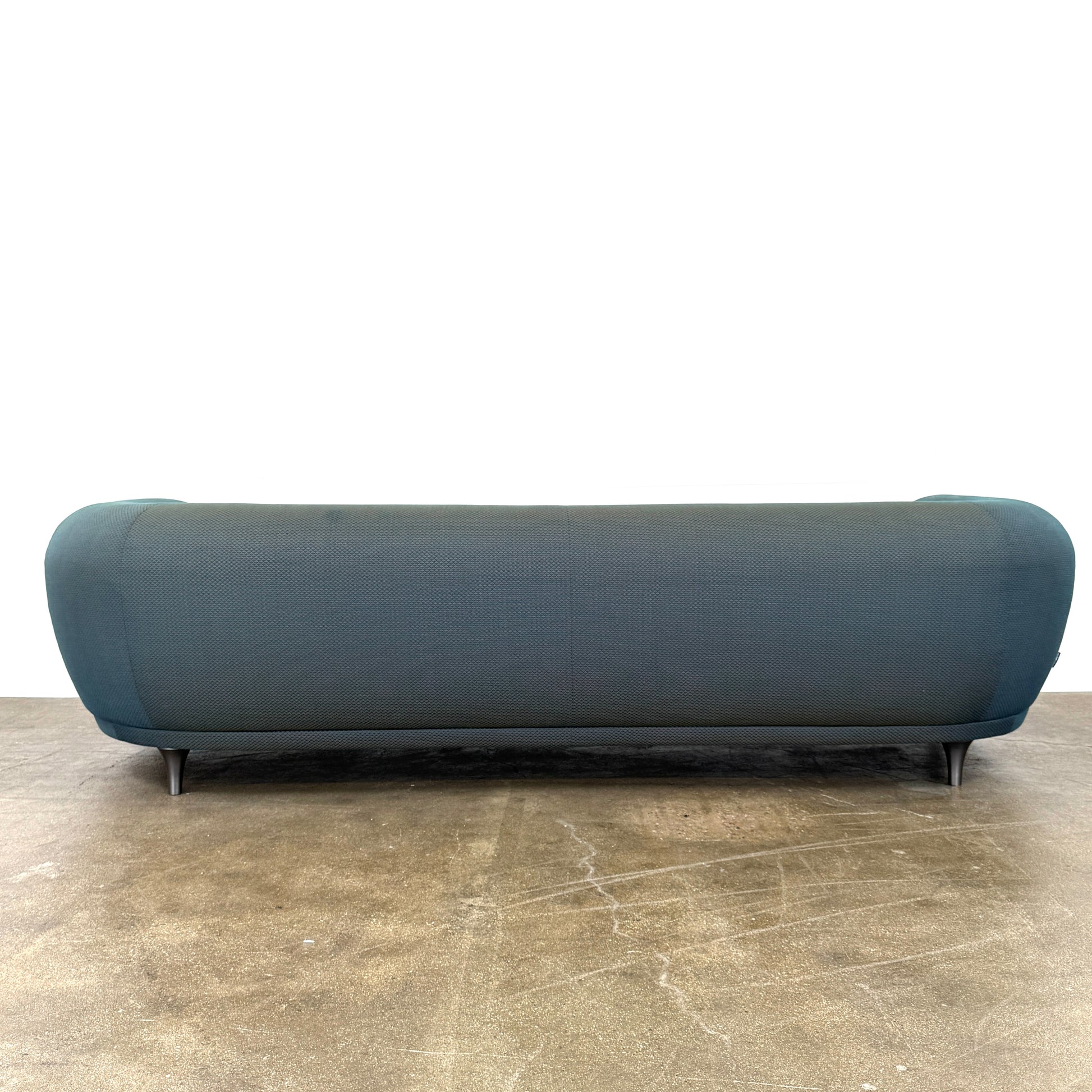 A Roche Bobois Montgolfiere sofa in dark gray, featuring rounded arms and black legs, from the Globe Trotter collection by Marcel Wanders, sits on a polished concrete floor against a plain white wall.
