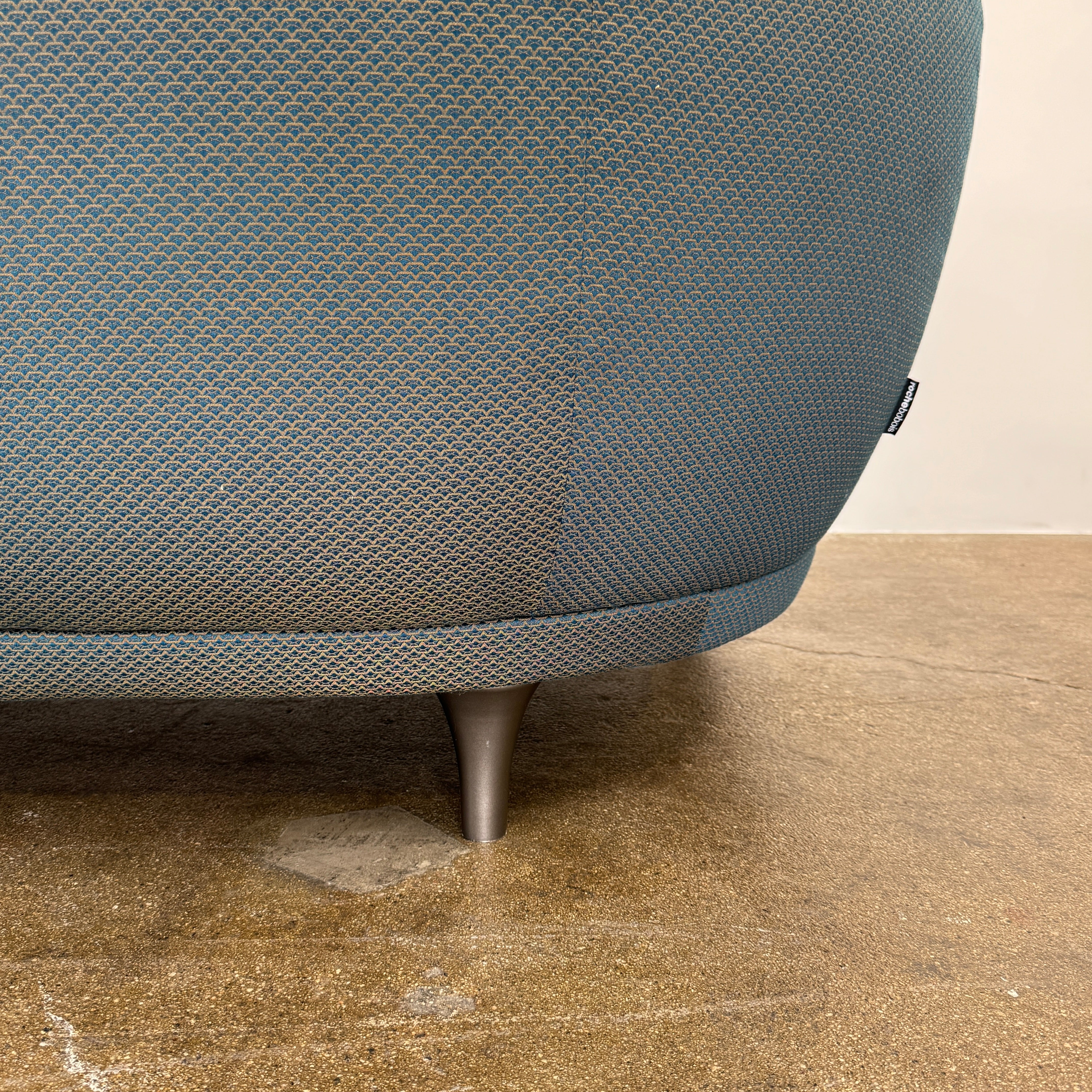 A Roche Bobois Montgolfiere sofa in dark gray, featuring rounded arms and black legs, from the Globe Trotter collection by Marcel Wanders, sits on a polished concrete floor against a plain white wall.