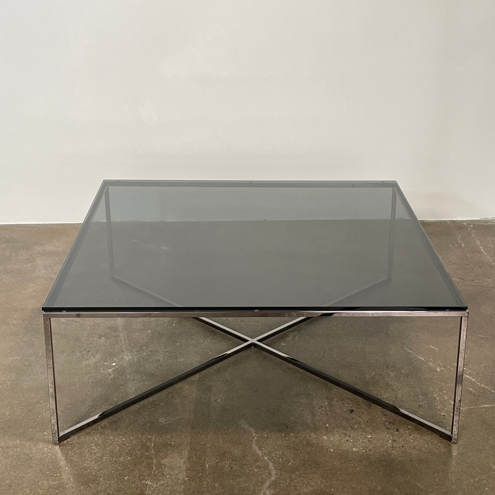 The Smoked Glass Coffee Table features a square design with a contemporary metal frame and crossed support bars, shown against a plain white background. Brand: Unknown.