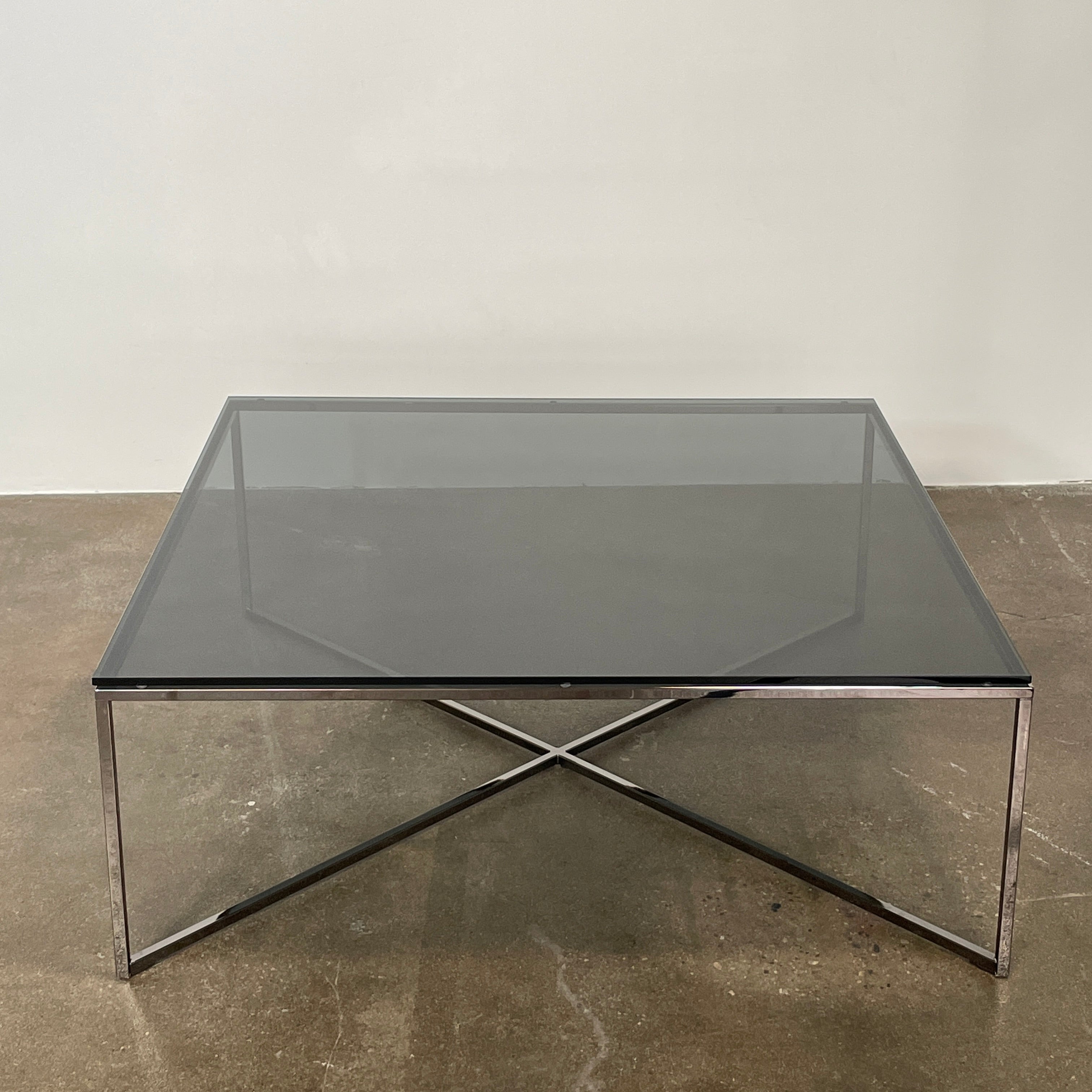 The Smoked Glass Coffee Table features a square design with a contemporary metal frame and crossed support bars, shown against a plain white background. Brand: Unknown.
