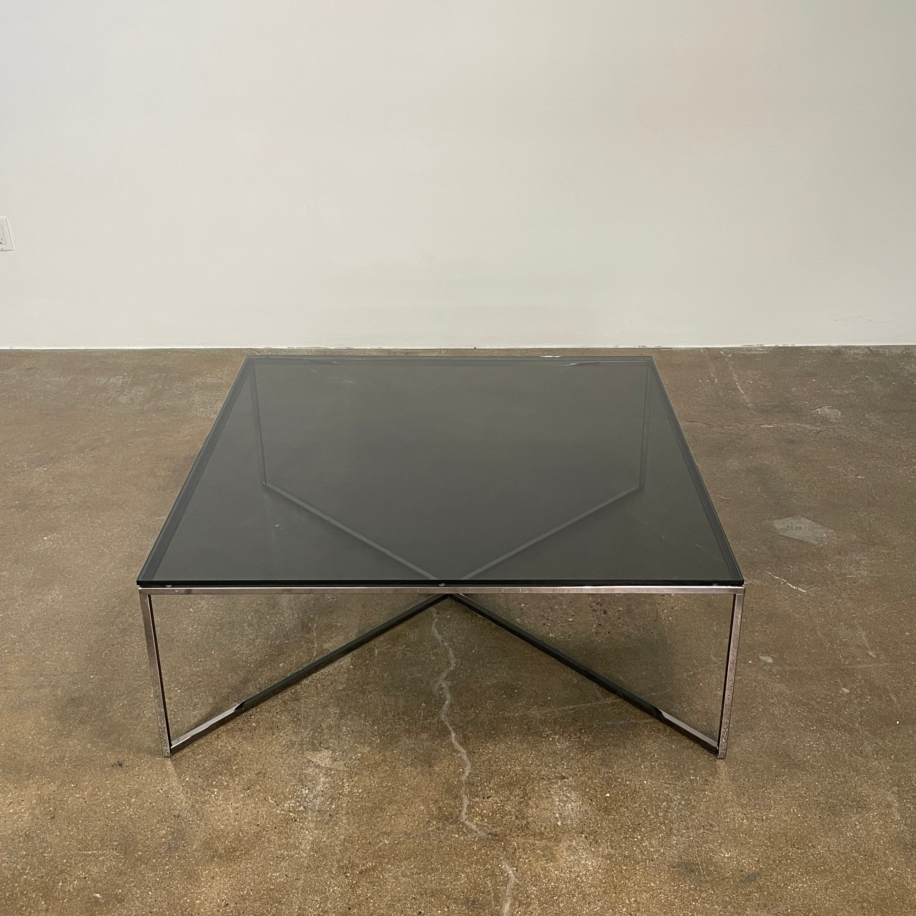 The Smoked Glass Coffee Table features a square design with a contemporary metal frame and crossed support bars, shown against a plain white background. Brand: Unknown.