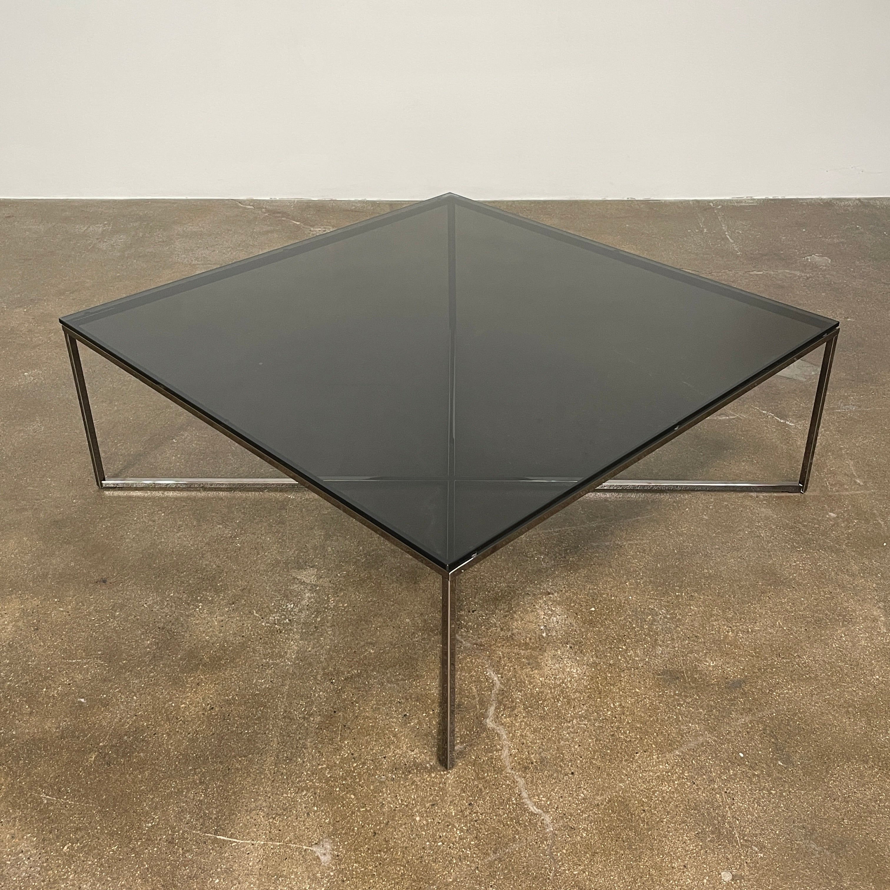 The Smoked Glass Coffee Table features a square design with a contemporary metal frame and crossed support bars, shown against a plain white background. Brand: Unknown.